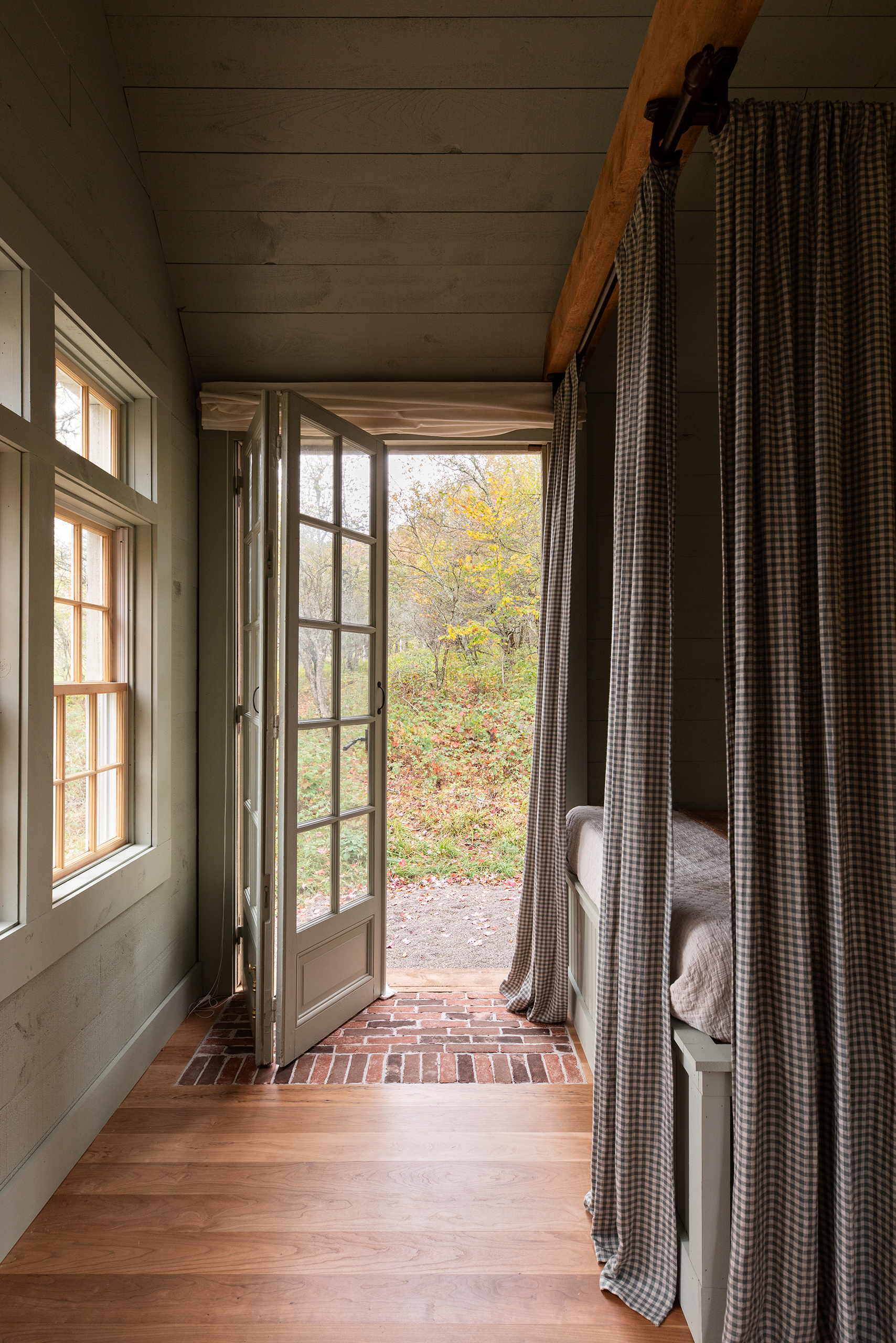 A photo looking out through an open door in the countryside. To the right is a bed with curtains fitted around it. The floorboards are bare and the walls and ceiling have been panelled.