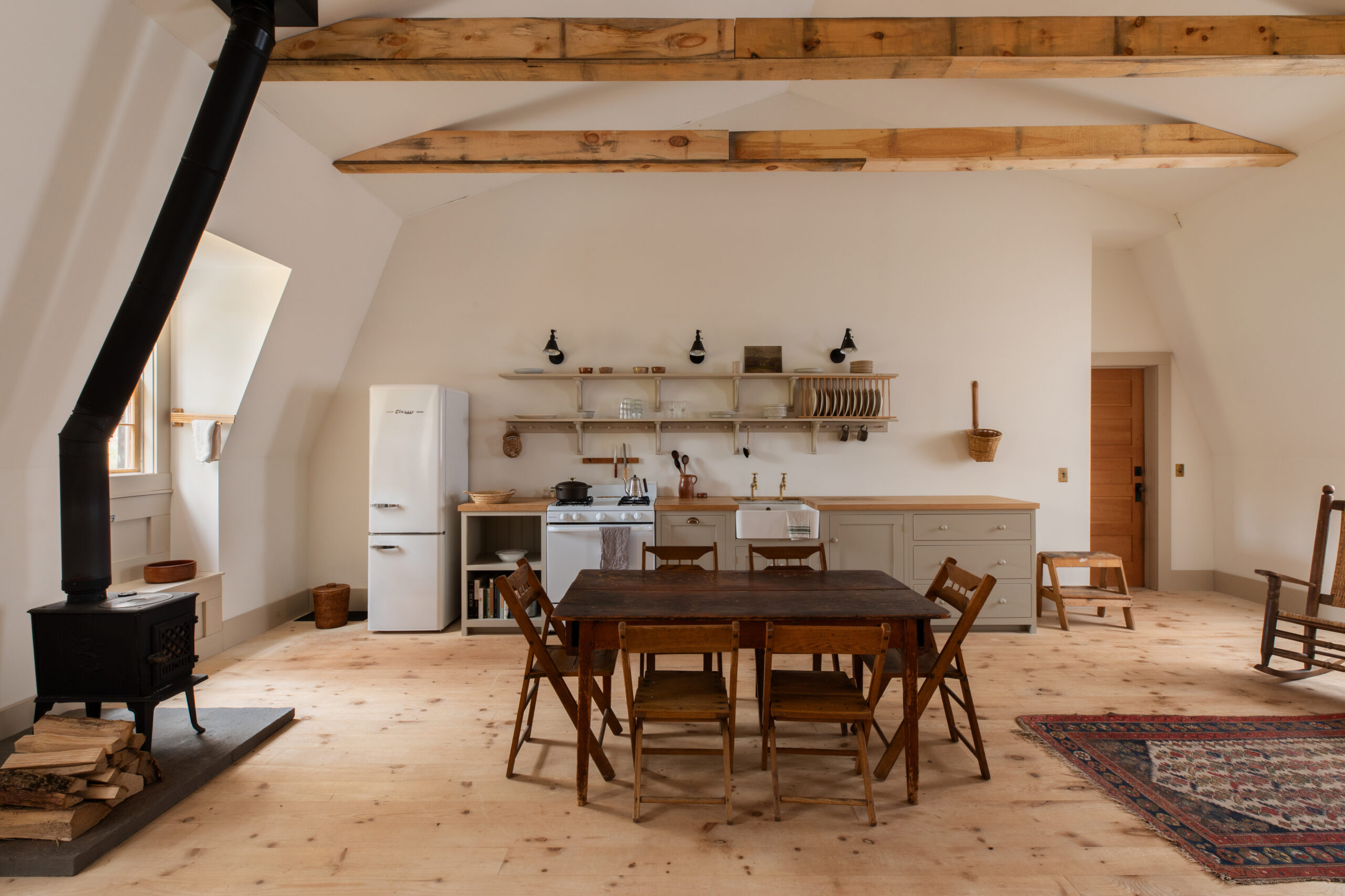 A landscape photo of a simple kitchen. Along the back wall is a run of shaker-style cupboard, it also houses a white oven and white fridge, as well as a ceramic sink. Above are two sets of open shelves. In front is a dining table with wooden chairs around it. To the left is a wooden burning stove.