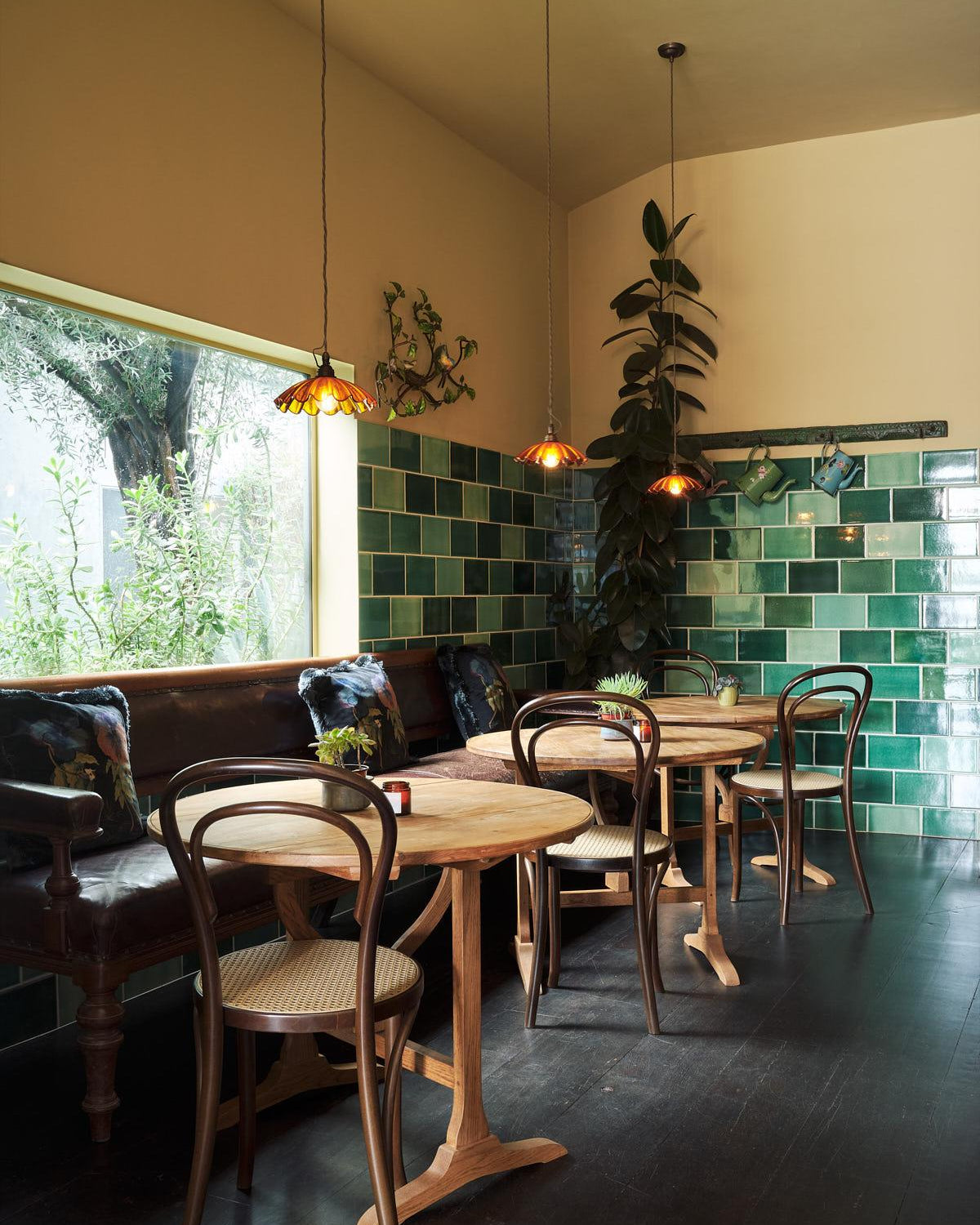 A cozy restaurant corner with wooden tables and chairs, a dark leather booth, green tiled walls, hanging pendant lights, and a large window looking out onto greenery.