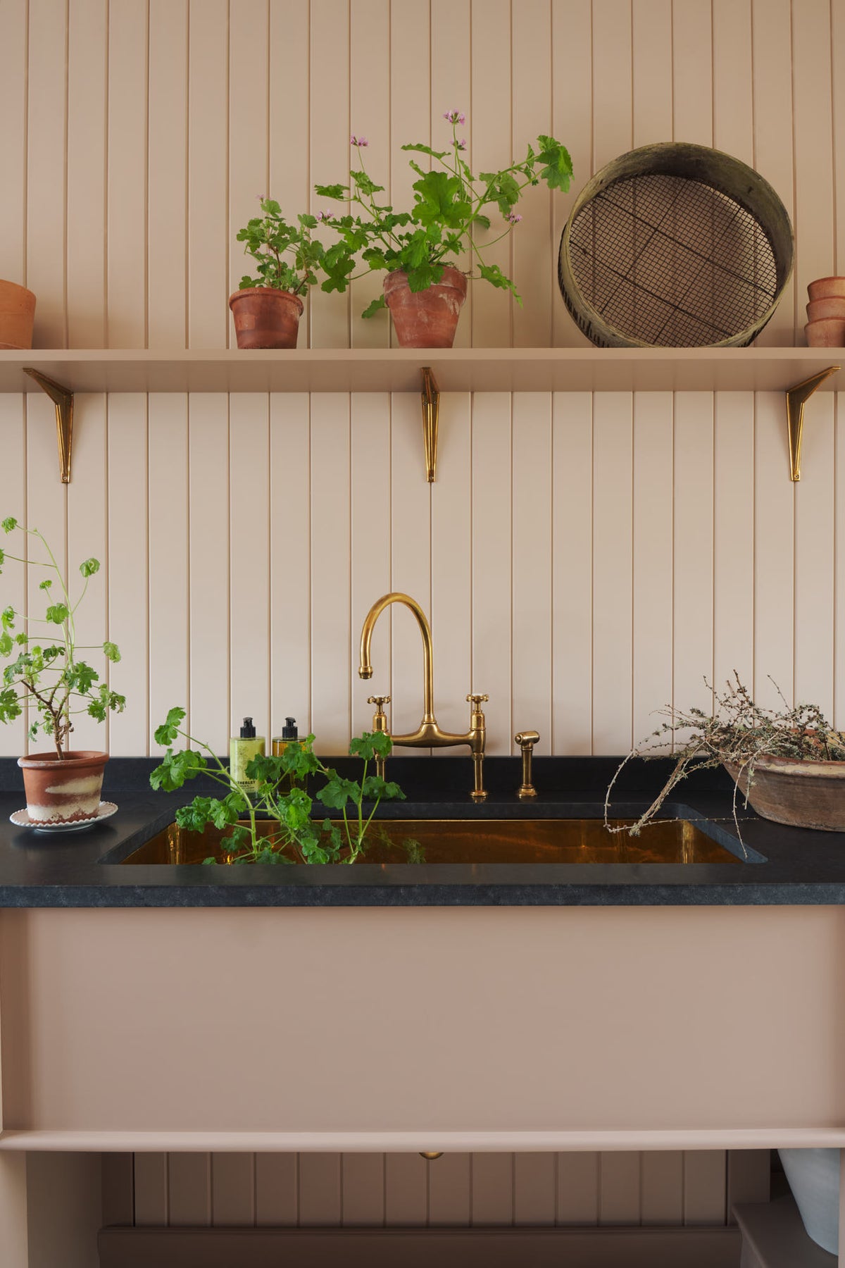 A closer look at the bohemian-chic, Princelet-pink Heirloom flower room featuring an Heirloom sink cabinet, an aged brass Ionian tap, large polished brass sink, and a long shelf. Beautifully decorated with artisan ceramics, vintage terracotta pots, and a collection of blooming house plants, it creates a soft and functional room for an aspiring florist.