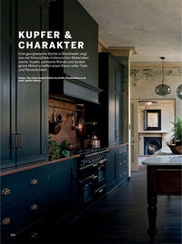 A page of a magazine which includes a full page image of a moody kitchen with dark green cupboards and copper worktops. The top left of the image has a block of white text. 