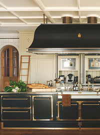 A page of a magazine, the whole page is taken up by a photo of a kitchen. In front is a large metal black island with matching extractor hood above. Behind is a run of tall wooden cabinetry painted in a soft beige. To the right is a wall of tiles and an arched doorway.