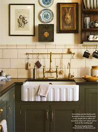 A page of a magazine, it shows a styled kitchen interior featuring a white farmhouse sink with a brass faucet, dark green cabinets, cream subway tile backsplash, and framed wall art above the sink.