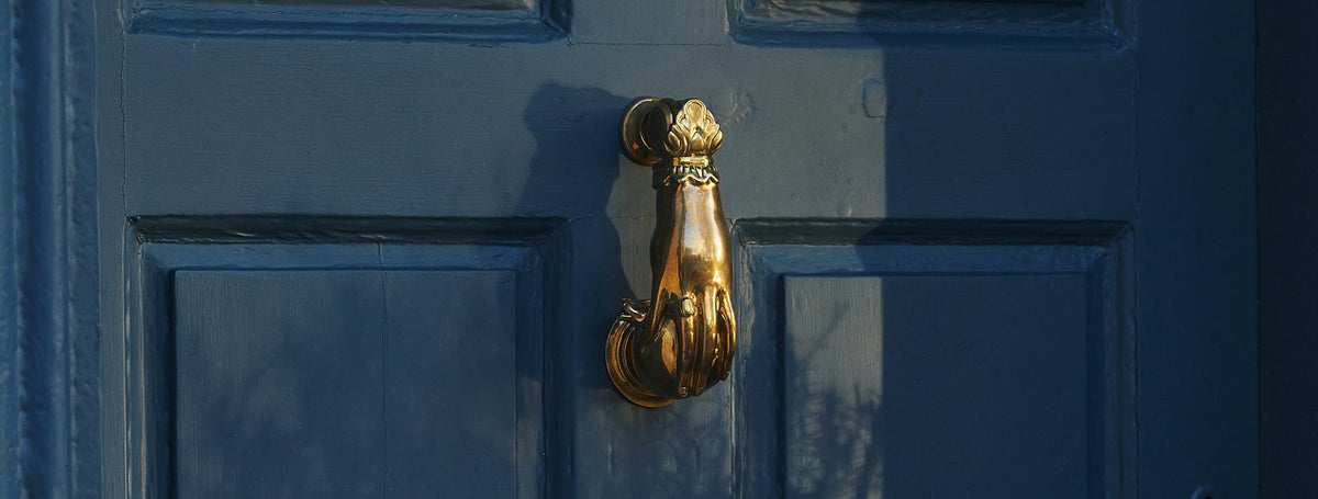 A decorative solid brass door knocker, shaped like a hand holding a pomegranate and fitted to a blue painted door.