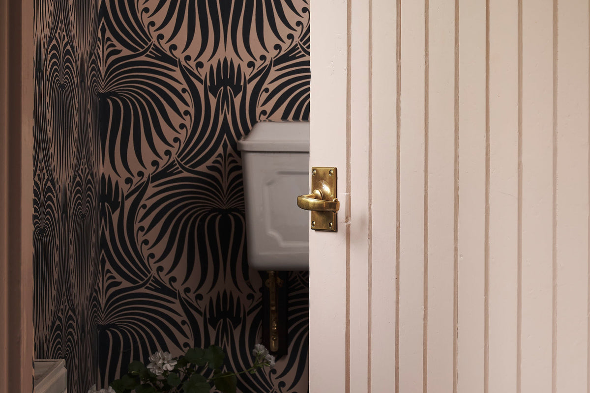 A close-up of the solid brass Otillie door handle with an Heirloom Brass finish. Promptly fixed on a Princelet pink door that is slightly opening into a small room with a pink wallpaper with black floral detail pattern.