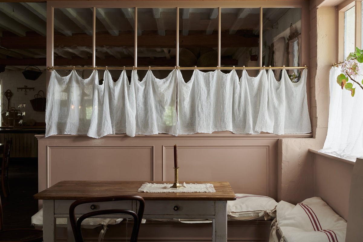Soft muslin fabric is gently draped from small brass clips on an Aged Brass Cafe Curtain Rail in front of a half-glazed partition wall painted in a dusty pink hue. In the foreground is an antique table, a glimpse of a deVOL Cafe Chair, and a single brass candle stick, creating a cosy seating area.