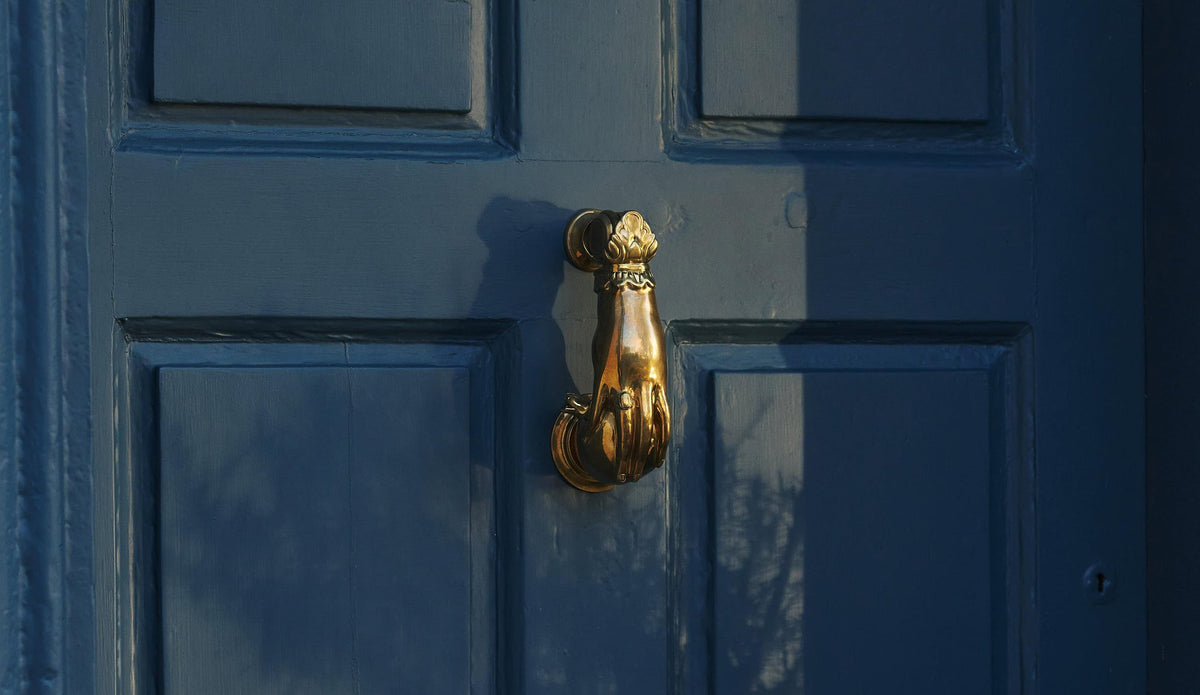 An oblique view of a decorative brass door knocker, shaped like a hand holding a pomegranate and fitted to a blue painted door.