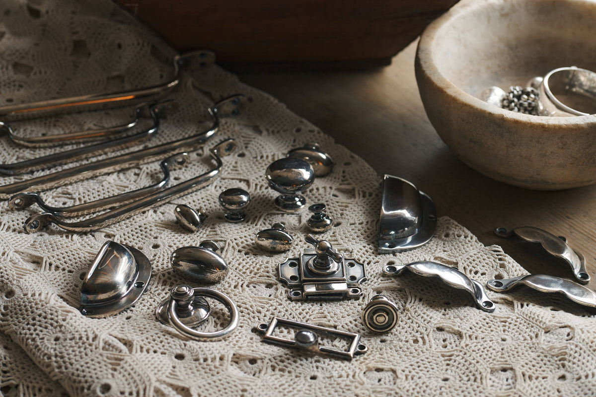 A collection of metal door knobs, handles and catches for cabinets and drawers in an Antique Silver finish, laid out on a delicate off-white knitted lace blanket.
