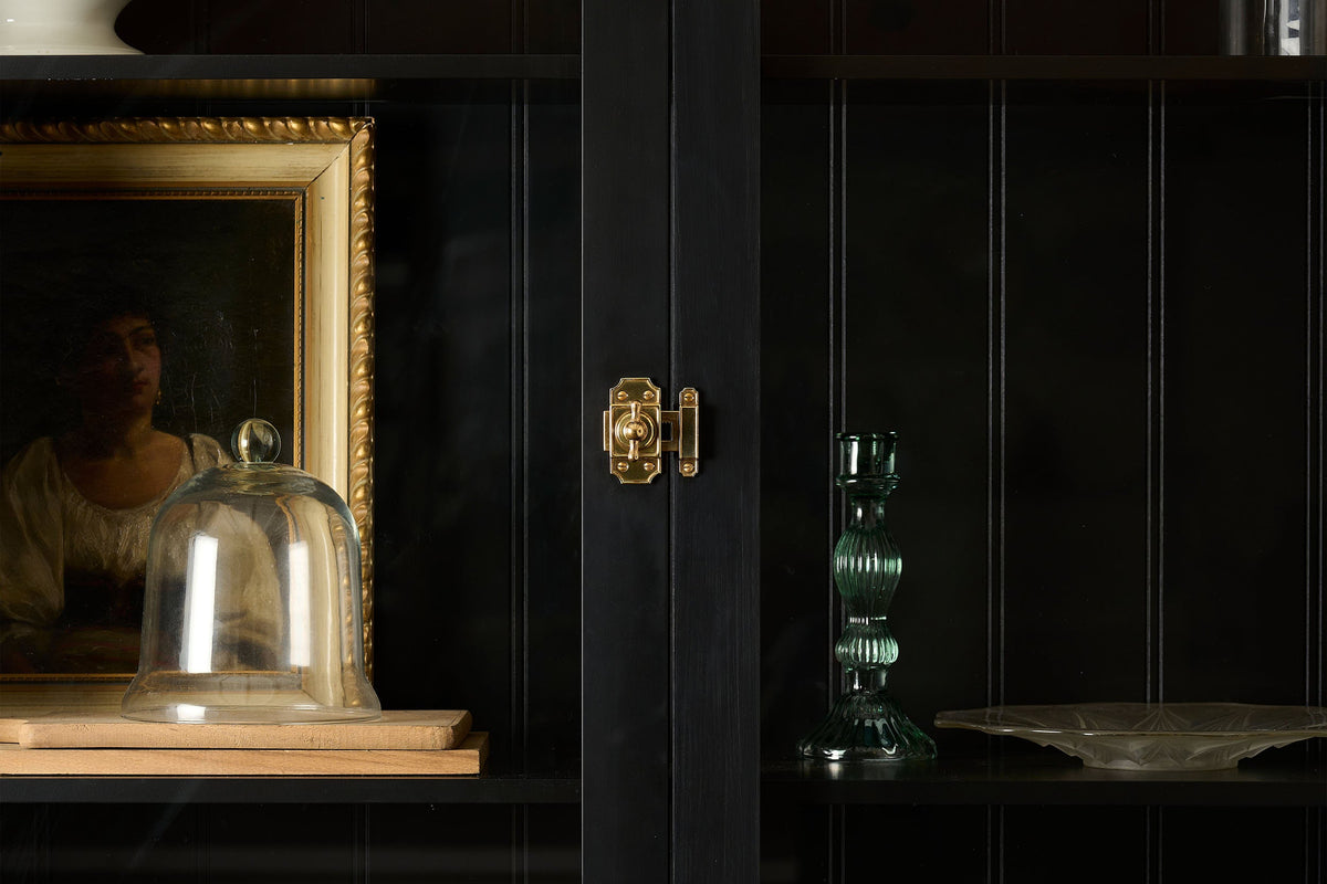 A straight-on view of a Victorian-inspired rack and pinion cupboard catch in a warm burnished brass finish, fitted to a tall glazed cupboard painted black inside and out.