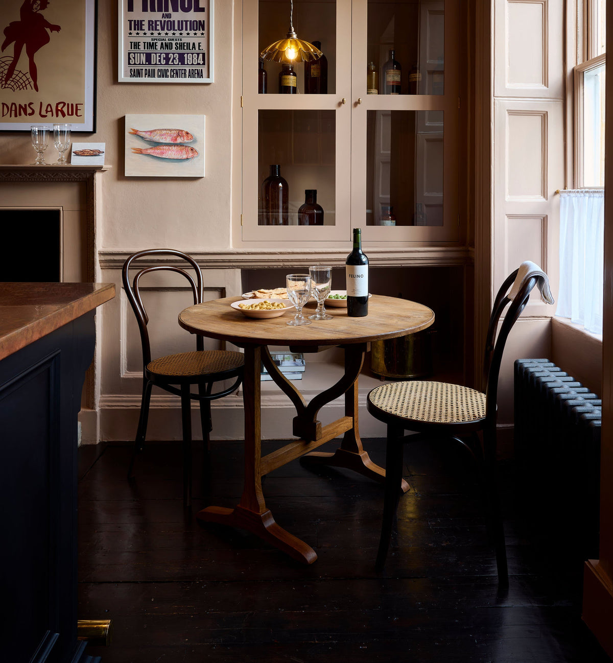 A quiet corner of a bold and dramatic Haberdasher’s kitchen features a deVOL petite vineyard table, Bentwood café chairs, and a petite frilly chartreuse light. Adorned with a delicious bottle of wine, drinking glasses, hors d’oeuvres, quirky framed illustrations and posters, and labelled glass jars, this space creates a local bistro bar atmosphere in Manhattan.