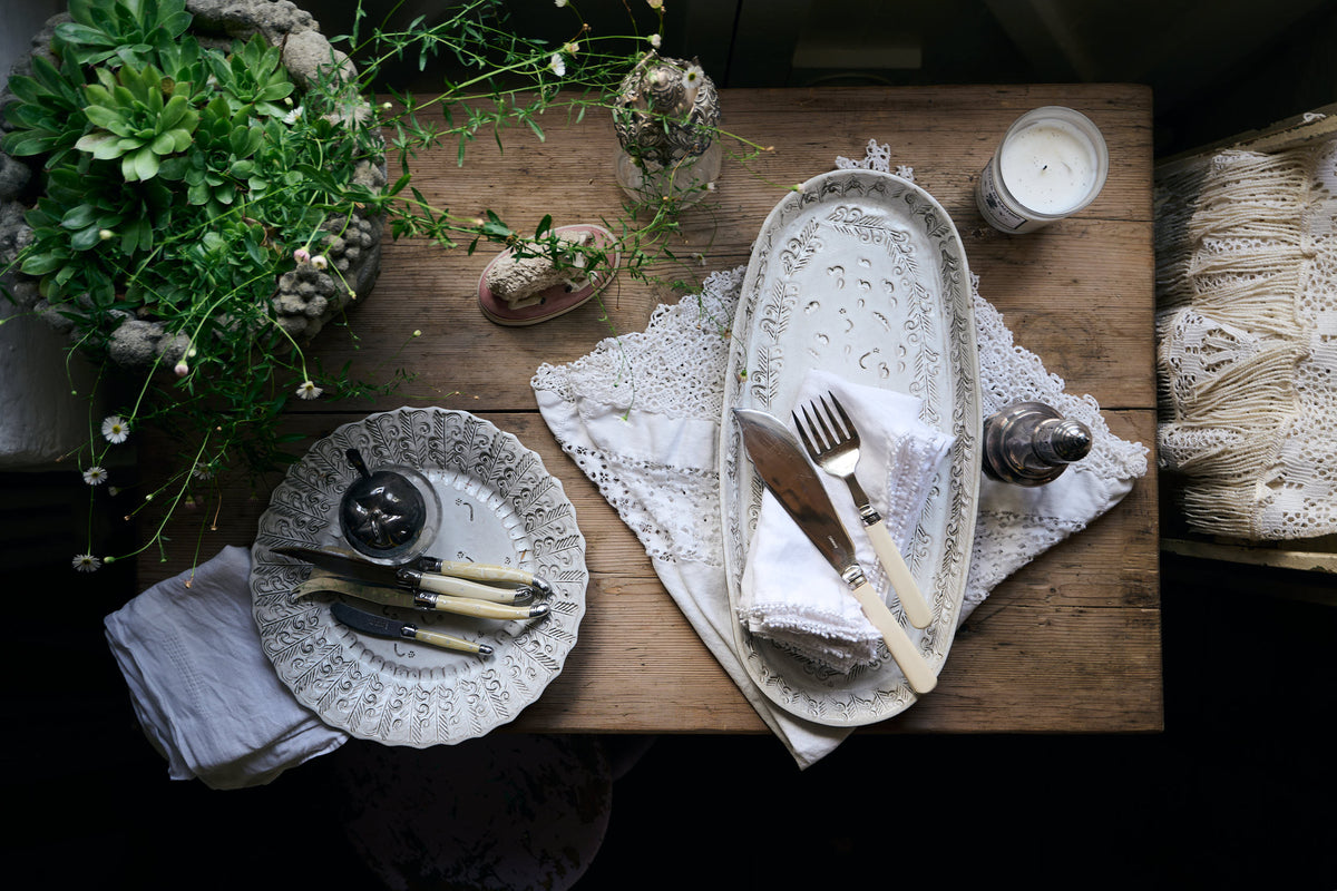An oval and round handmade bohemian-style white ceramic platter with delicate impressions and surface patterns, complimented perfectly by vintage silverware, white linens and a rustic pine tabletop.