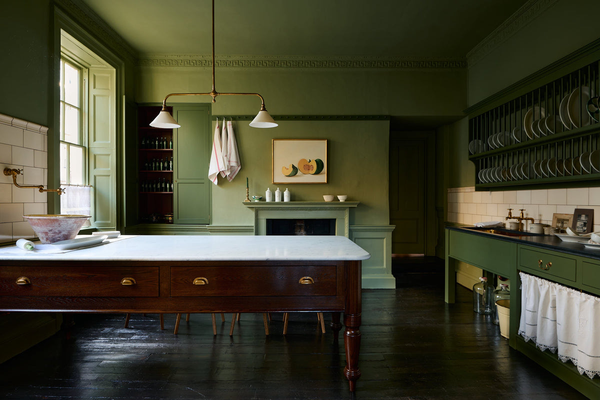 A quaint and stylish Wilkes green Heirloom kitchen features a sink cabinet with a café rail, white lace curtains, a polished brass sink, aged brass Mayan taps, a large plate rack, and a dairy table. Adorned with an aged brass pot filler, artisan ceramics, Heirloom gaselier lights, cream subway splashback tiles, and a golden framed sliced green pumpkin, this kitchen creates a luxurious dining and social space.