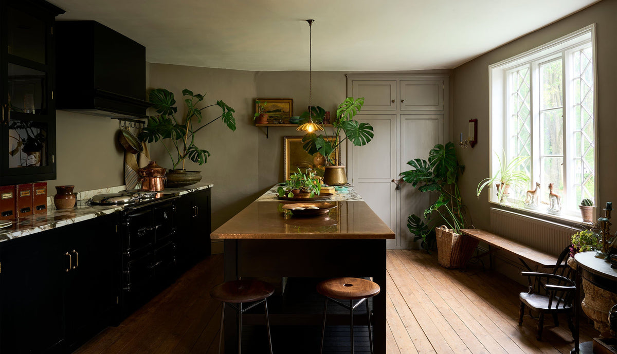 An exotic and glamorous kitchen with black painted Shaker cupboards, warm grey walls with a fitted Classic English pantry, opulent copper and quartzite worktops, original worn floorboards and beautiful leaded windows. The room is brought to life by the green and gold accents of three large monstera plants, a dazzling frilly glass chartreuse pendant light, antique oil paintings and other vintage oddities.