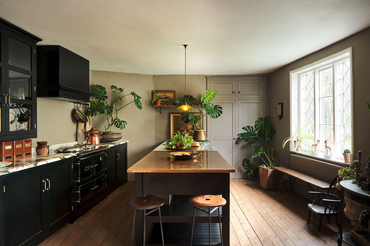 An exotic and glamorous kitchen with black painted Shaker cupboards, warm grey walls with a fitted Classic English pantry, opulent copper and quartzite worktops, original worn floorboards and beautiful leaded windows. The room is brought to life by the green and gold accents of three large monstera plants, a dazzling frilly glass chartreuse pendant light, antique oil paintings and other vintage oddities.