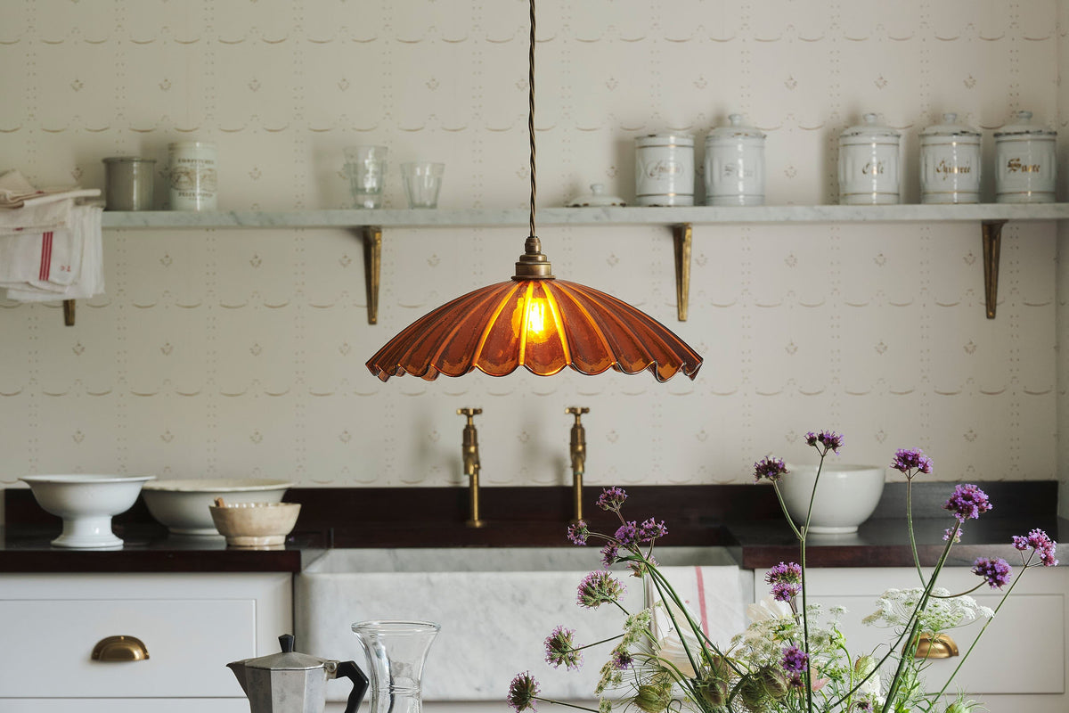 A close-up view of a grand frilly amber glass pendant light with brass fittings and brown twisted fabric flex cord. The light is on and shining through the translucent glass.