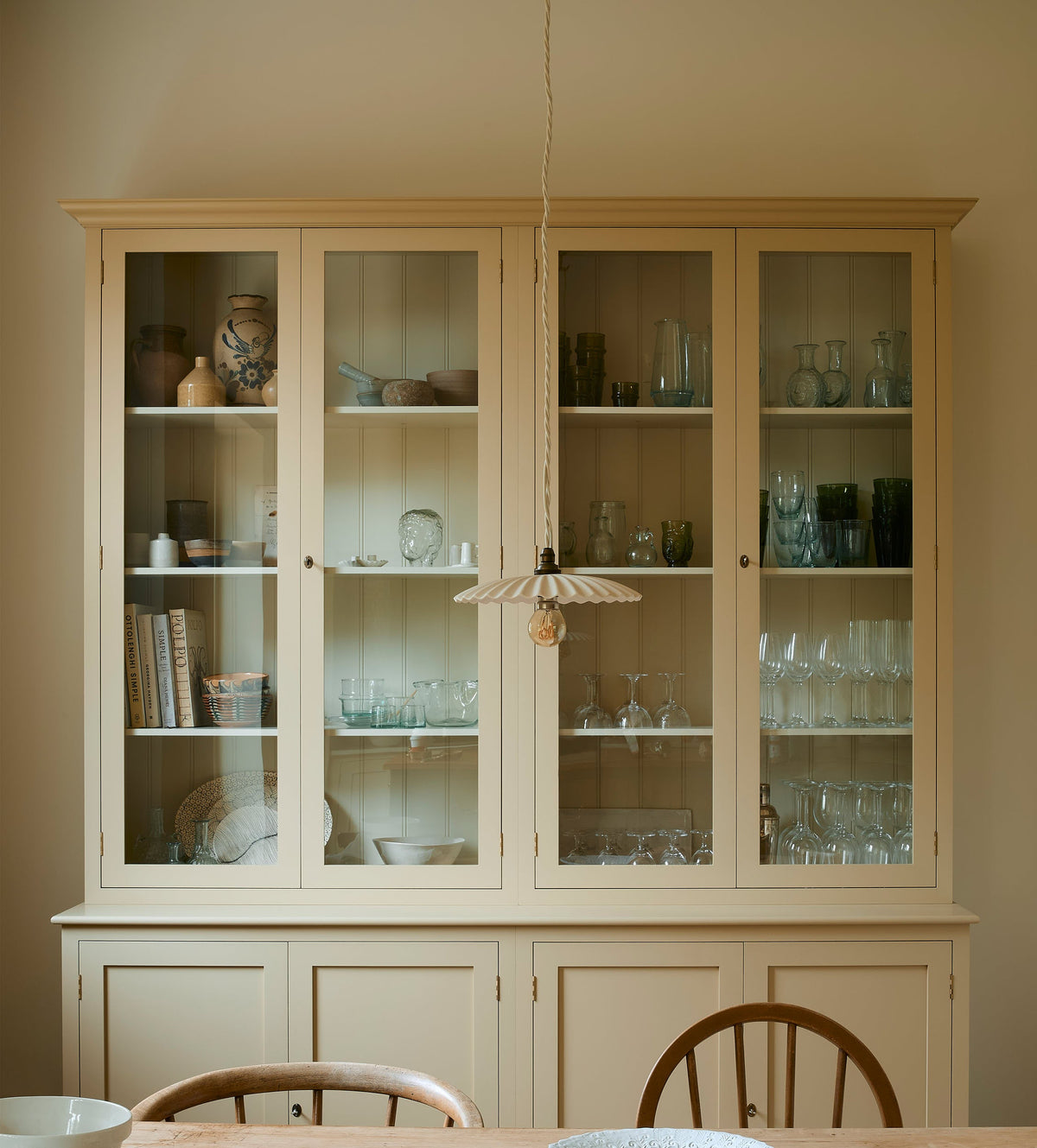  A cream, glazed pantry cupboard stands proudly at the back of a shaker kitchen. In the foreground is an old pine table and antique stick-back chairs, styled with vintage lace, an antique silver spoon and deVOL’s heirloom round platter. A delicate frilly pendant light hangs above the table.