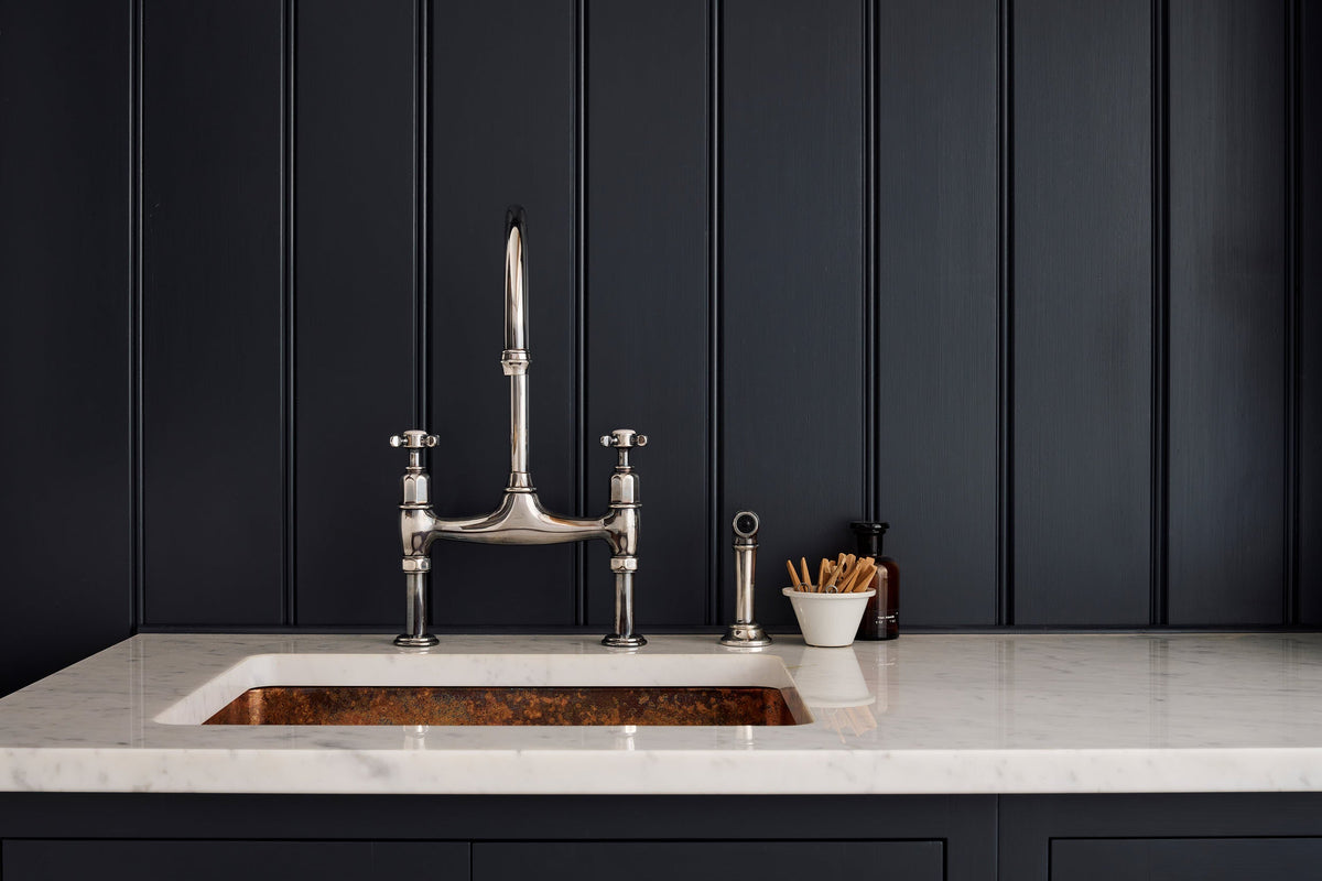 A close-up of a simple pantry blue shaker kitchen's sink run featuring a n antique silver ionian tap, marble countertop and a copper sink.