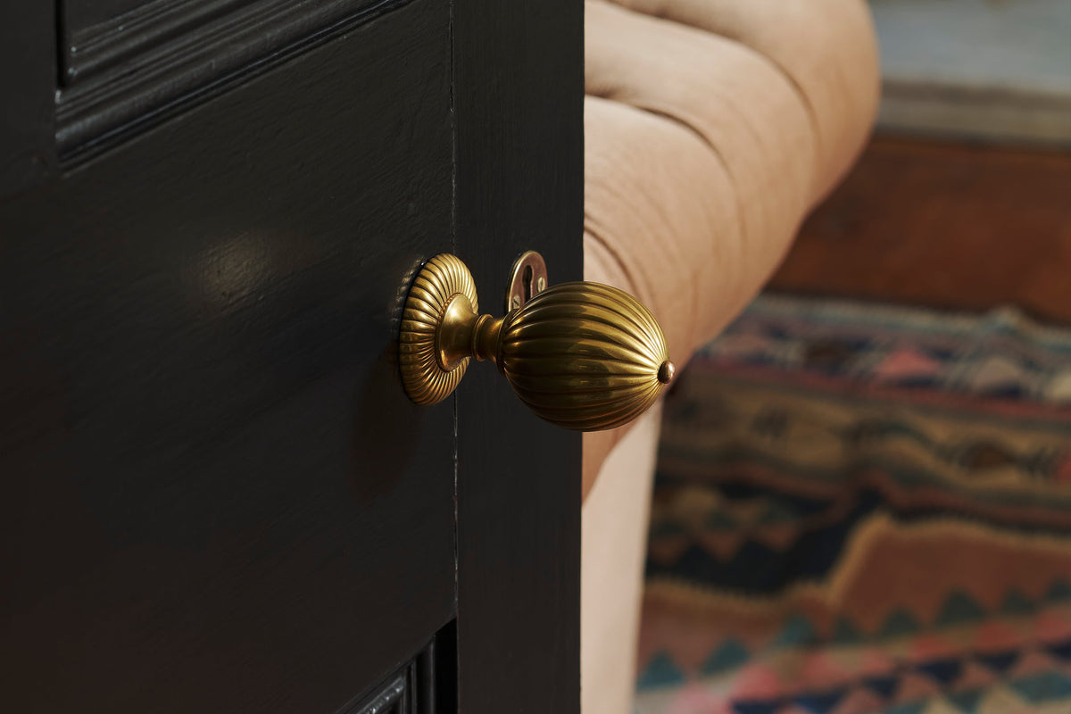 A close-up of the solid brass Cecily doorknob with an Heirloom Brass finish. Promptly fixed on a black door, highlighting the rimmed design with a bronze button detail.