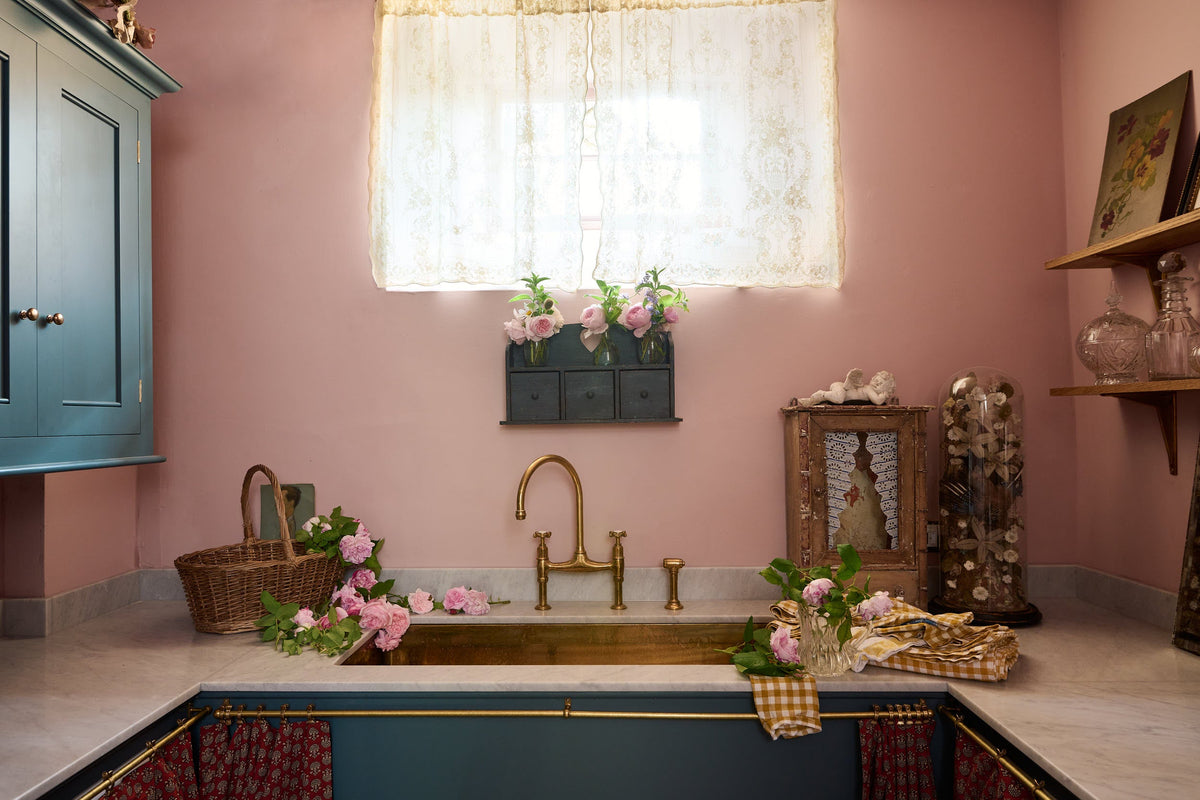 A small and practical Heirloom laundry room features a sink cabinet, a polished brass sink, an aged brass Ionian tap, a curtain rail with crest-patterned red curtains, wall cupboards, and shelves. Adorned with eclectic vintage items, pink peonies, and a small wicker basket, this space creates a romantic and functional atmosphere full of personality.