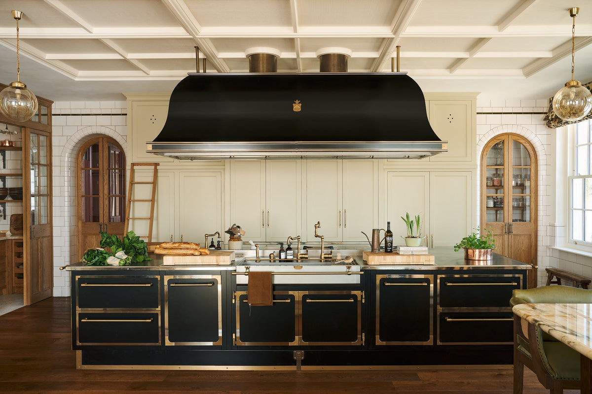 A grand room with high panelled ceiling, traditional glossy white tiled walls with curved archways on either side of a huge bank of creamy mushroom fitted full-height Classic cupboards with a polished oak ladder. An elaborate black with gold accent kitchen island and canopy sits in the middle of the space with an array of articulated brass sink taps around a wide and shallow sink.