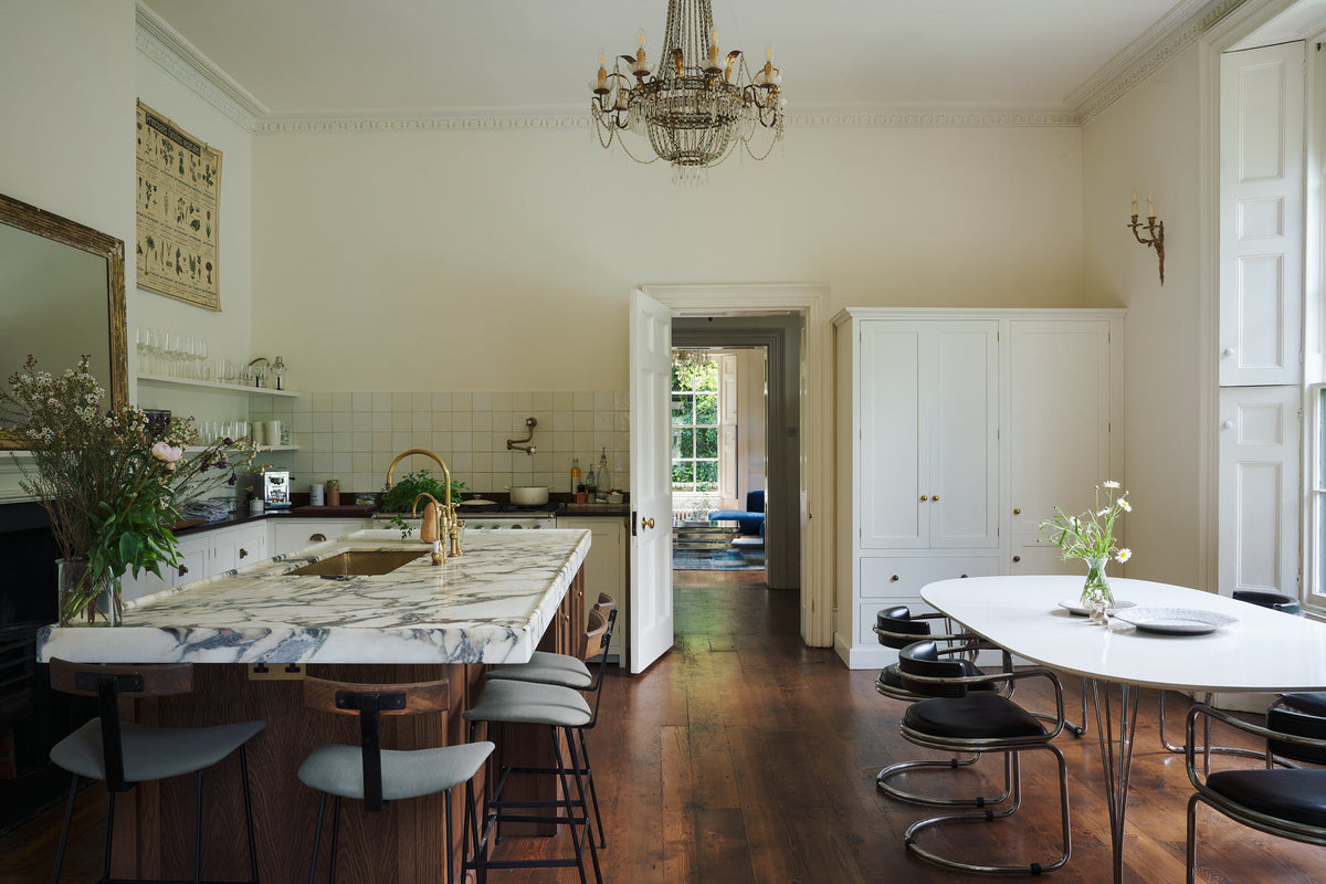 A classic English kitchen in white linen features white and cream splashback tiles, a cooker with a polished sapelle worktop, and a kitchen island topped with Arabescato marble. It includes a polished brass sink, an aged brass Ionian tap, a scientific illustration of vegetable species, two glasses of local wildflowers, and a metal-and-crystal chandelier.