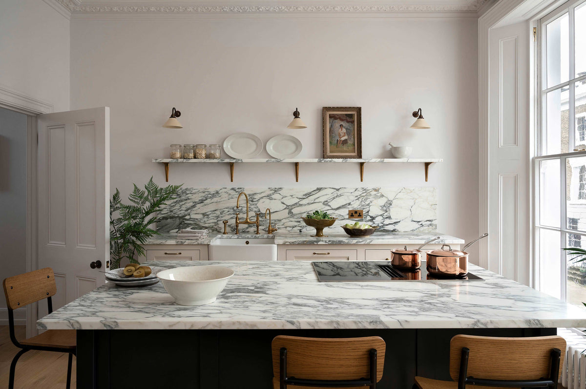 A humble yet elegant off-white Classic English kitchen features a sink run with honed Arabescato marble worktop and splashback, a pantry blue kitchen island with a cooker, mini creamware wall lights, and plumber’s stools. Adorned with artisan ceramics, brass pans, a vintage painting, fresh greens, and grains, creating an intimate social and practical kitchen space.