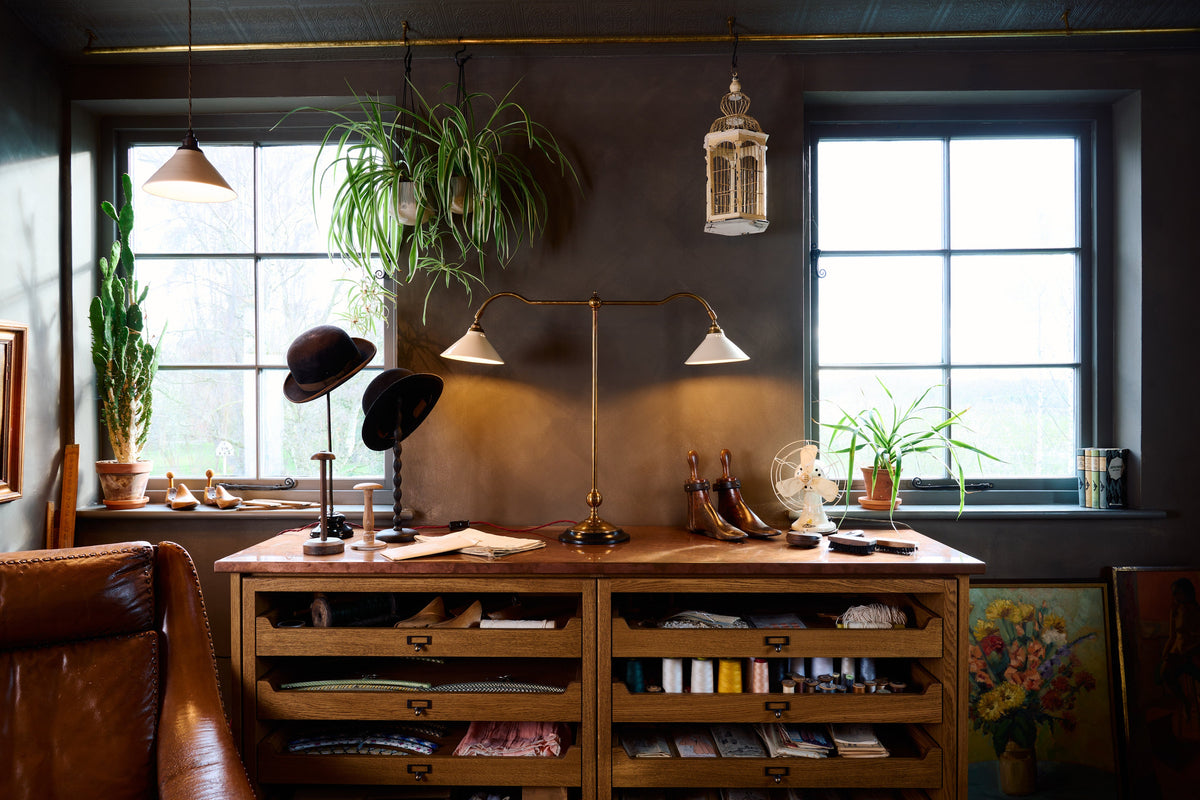 A straight-on view of a Victorian-inspired desk lamp with a solid brass foot, stand, two curved arms and two small cream glazed ceramic light shades on top of haberdasher's cupboards.