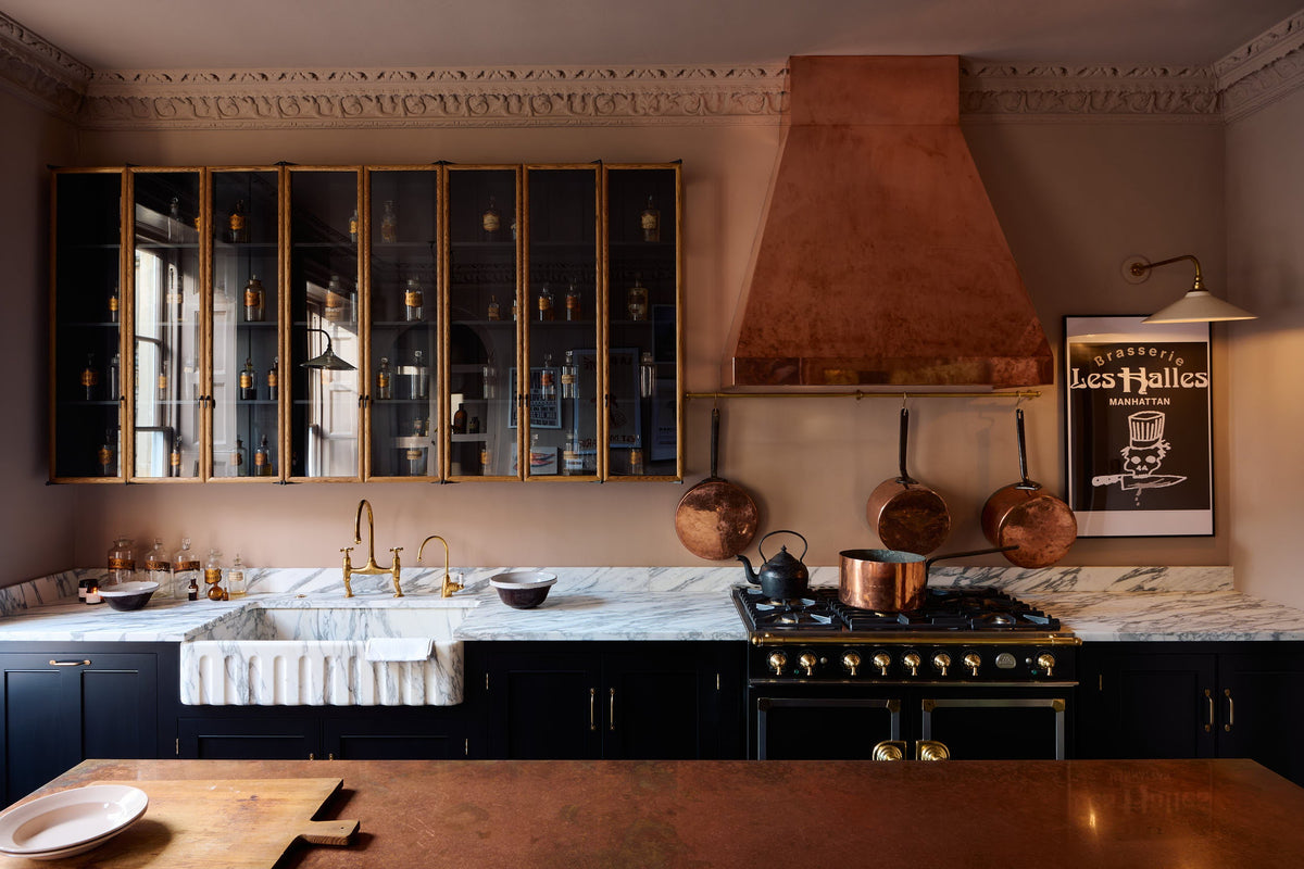 A bold and dramatic Habersasher’s kitchen with a sink and cooker run, an Arabescato worktop and single fluted sink, an aged brass Ionian tap, a glazed wall cupboard, a large copper canopy, and a kitchen island with a copper worktop. Adorned with copper pans, a milk glass task light, artisan ceramics, labelled glass jars, and a quirky framed illustration, creating a local bristo bar in Manhattan space.