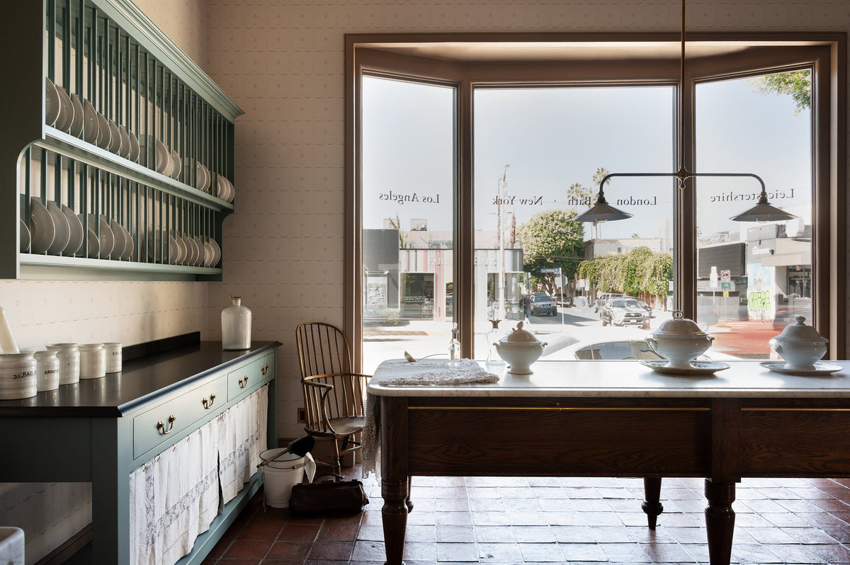 A grand and spacious Heirloom kitchen with Trinity blue open cabinets and plate racks, Heirloom Dairy Tables, an Heirloom Gaselier light reaching all the way to the top of the high ceiling and rustic Square Terracotta tiles. Beautifully decorated with a spectacular chandelier, artisan ceramics, and plates of green pears.