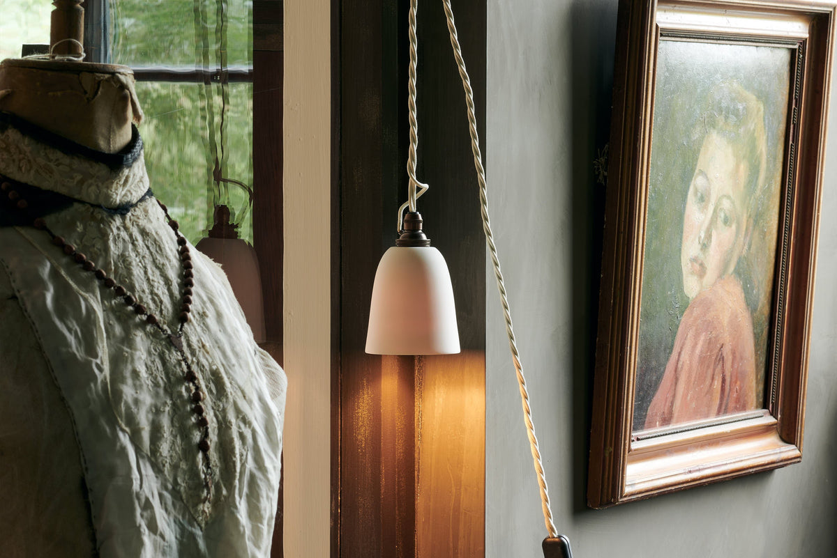 A close-up of a plug-in porcelain beaker light, hung up on the wall beside a vintage painting and tailor's dummy.