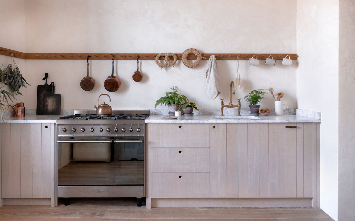 A light contemporary kitchen with creamy textured walls, rough-sawn beech slatted cupboard doors, an aged brass tap and a span of wooden pegs for hanging mugs, pans and utensils above a mottled white marble worktop. The styling is simple, Scandinavian and slightly rustic, with small potted ferns and woven rope trivets, and a built-in oven.