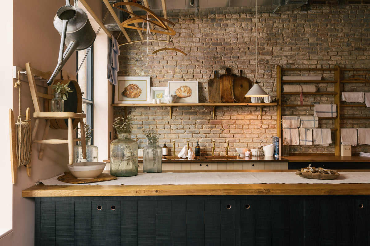 A front view of a rustic Sebastian Cox freestanding island with a solid oak worktop and sink run, featuring a copper worktop, two aged copper single sinks, and two sets of aged brass Mayan taps, complemented by café curtain tails. The cabinets highlight the raw ingrains and unique characteristics of the wood. Even when stained black, they harmonise beautifully with the love of natural materials.