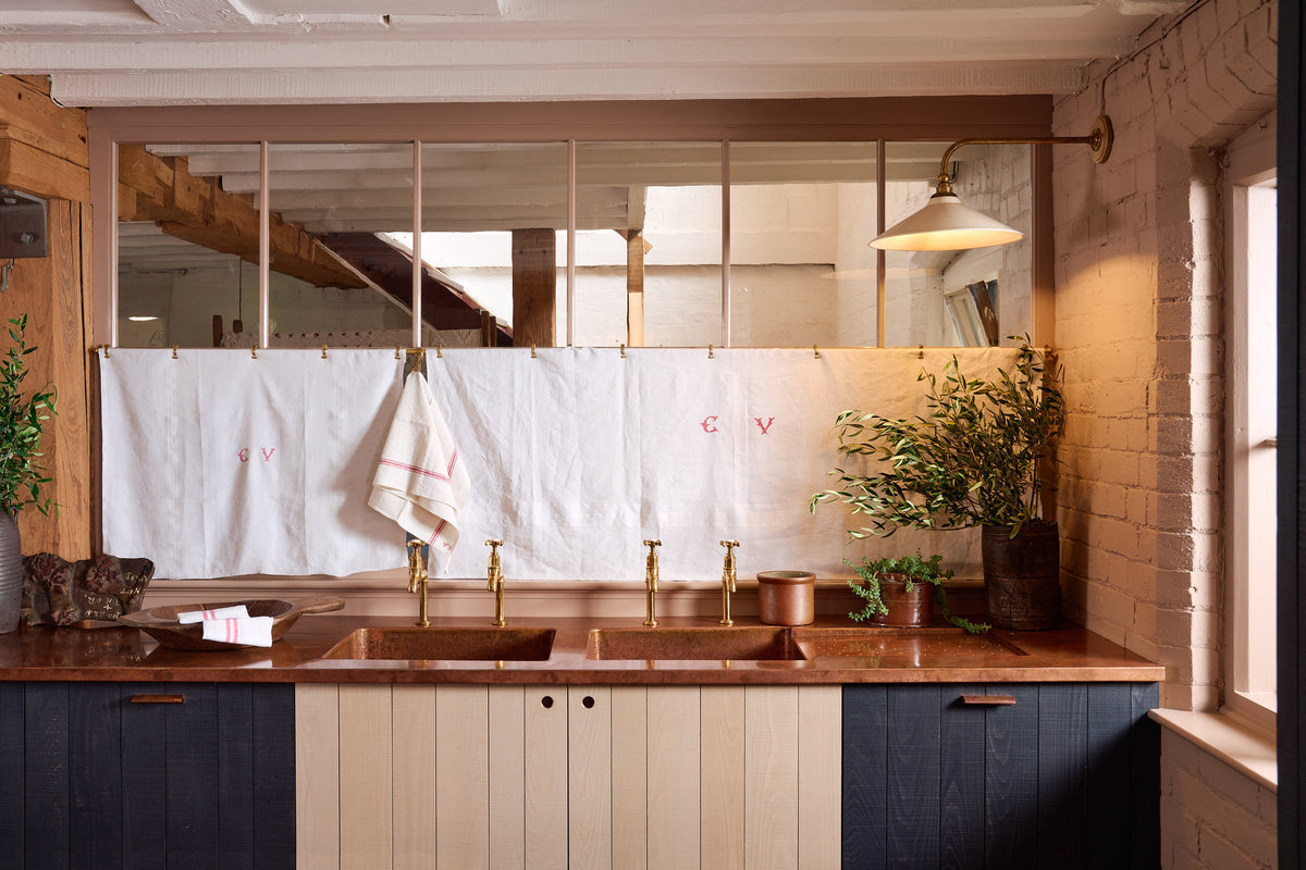 A warm and stylish Sebastian Cox kitchen features a sink run with two single copper sinks, aged brass Mayan taps, glazed partitions, and a copper worktop. Adorned with an aged brass café curtain rail, a milk glass task light, artisan ceramics, and vintage wooden items, this kitchen creates a humble space that celebrates a love of simple and natural materials.