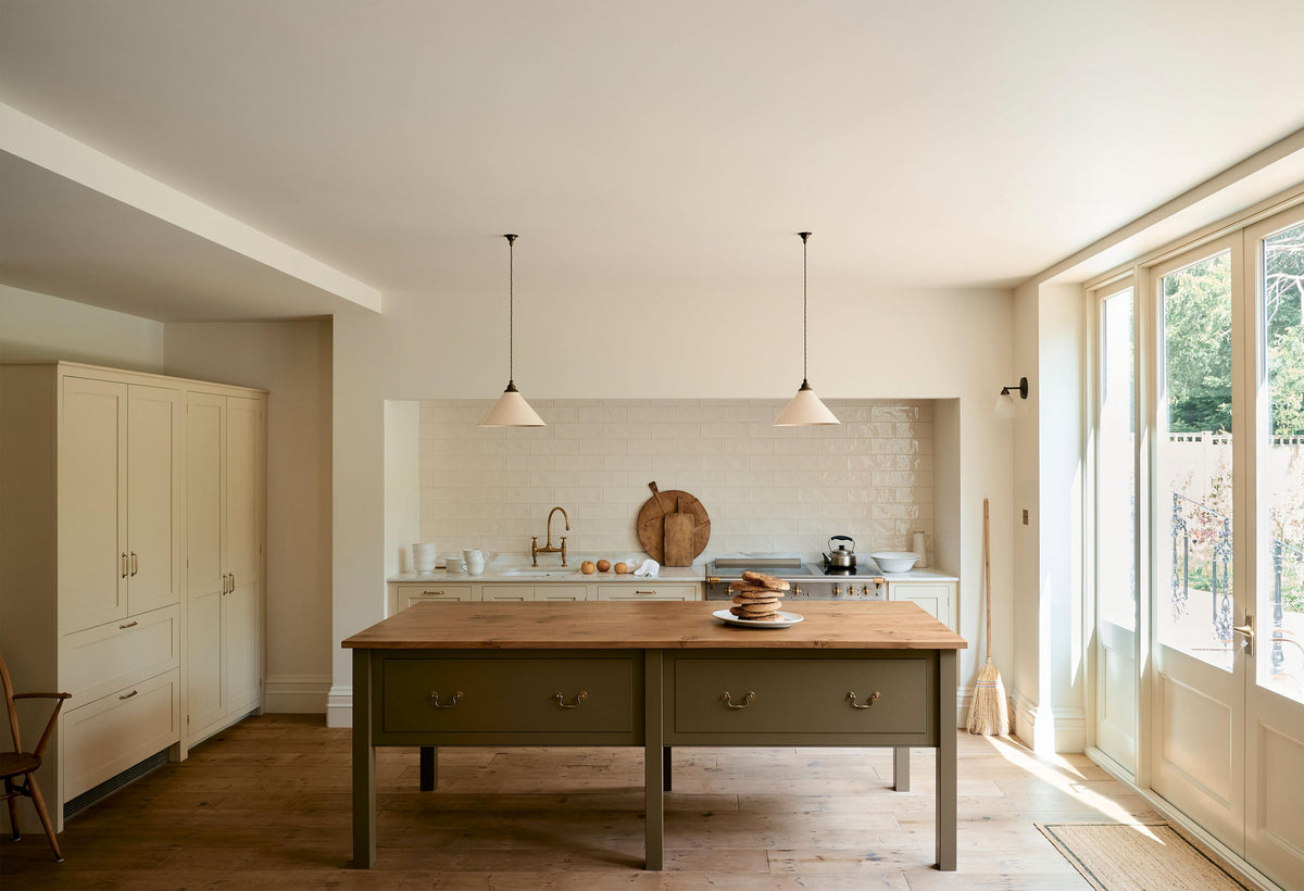 A light, clean, and simple shaker kitchen features a sink and cooker run nestled in a chimney breast, cream porcelain splashback tiles, wooden floorboards, and white walls. Cream cabinets complement an olive green Georgian-style prep table with deep drawers and a rustic oak worktop at the room’s centre, illuminated by two elegant creamware pendant lights.
