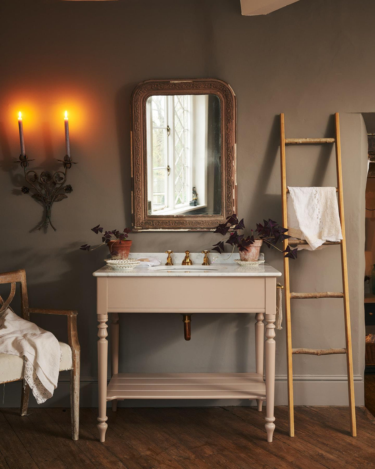 A straight-on view of a dusty-pink Victorian-inspired washstand with brass mixer taps in a greyish-brown painted room. The shot is atmospheric and moody, with two lit candles casting a subtle glow and light from an adjacent window illuminating the brass accents. | Large Princelet Pink shown TEST