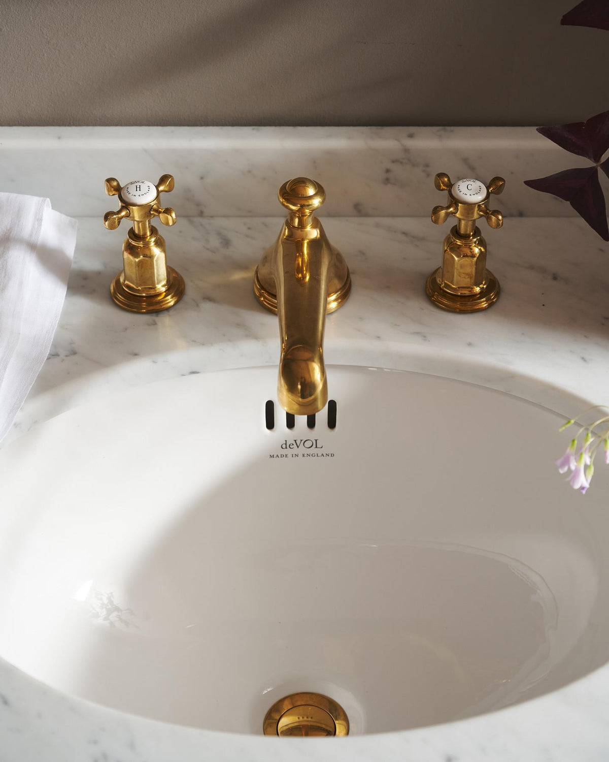 A close-up oblique view of the crosshead handle of an aged brass mixer tap for a bathroom basin sink. The letter 'H' is in the centre of the handle top, surrounded by the words 'deVOL, Made in England'. TEST