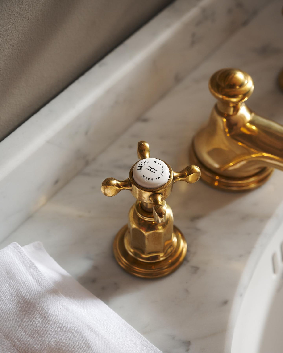 A close-up oblique view of the crosshead handle of an aged brass mixer tap for a bathroom basin sink. The letter 'H' is in the centre of the handle top, surrounded by the words 'deVOL, Made in England'. TEST