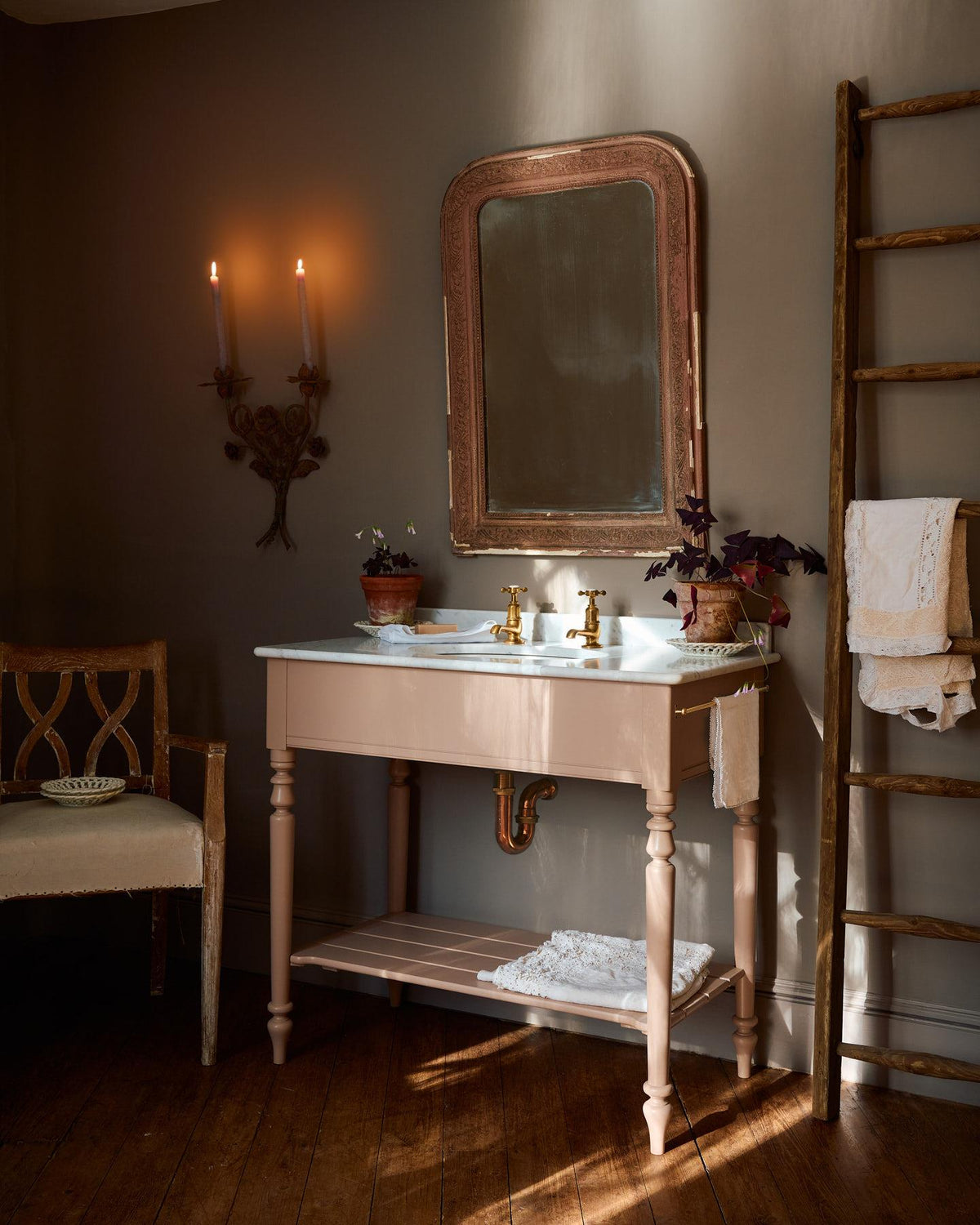 An oblique view of a dusty-pink Victorian-inspired washstand with brass pillar taps in a greyish-brown painted room. The shot is atmospheric and moody, with two lit candles casting a subtle glow and light from an adjacent window illuminating the brass accents. | Large Princelet Pink shown TEST