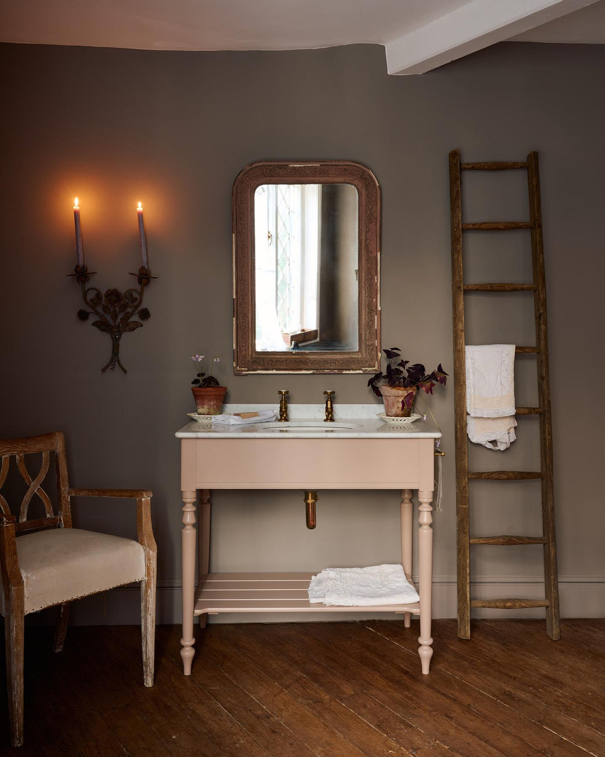 A straight-on view of a dusty-pink Victorian-inspired washstand with brass pillar taps in a greyish-brown painted room. The shot is atmospheric and moody, with two lit candles casting a subtle glow and light from an adjacent window illuminating the brass accents. | Large Princelet Pink shown TEST