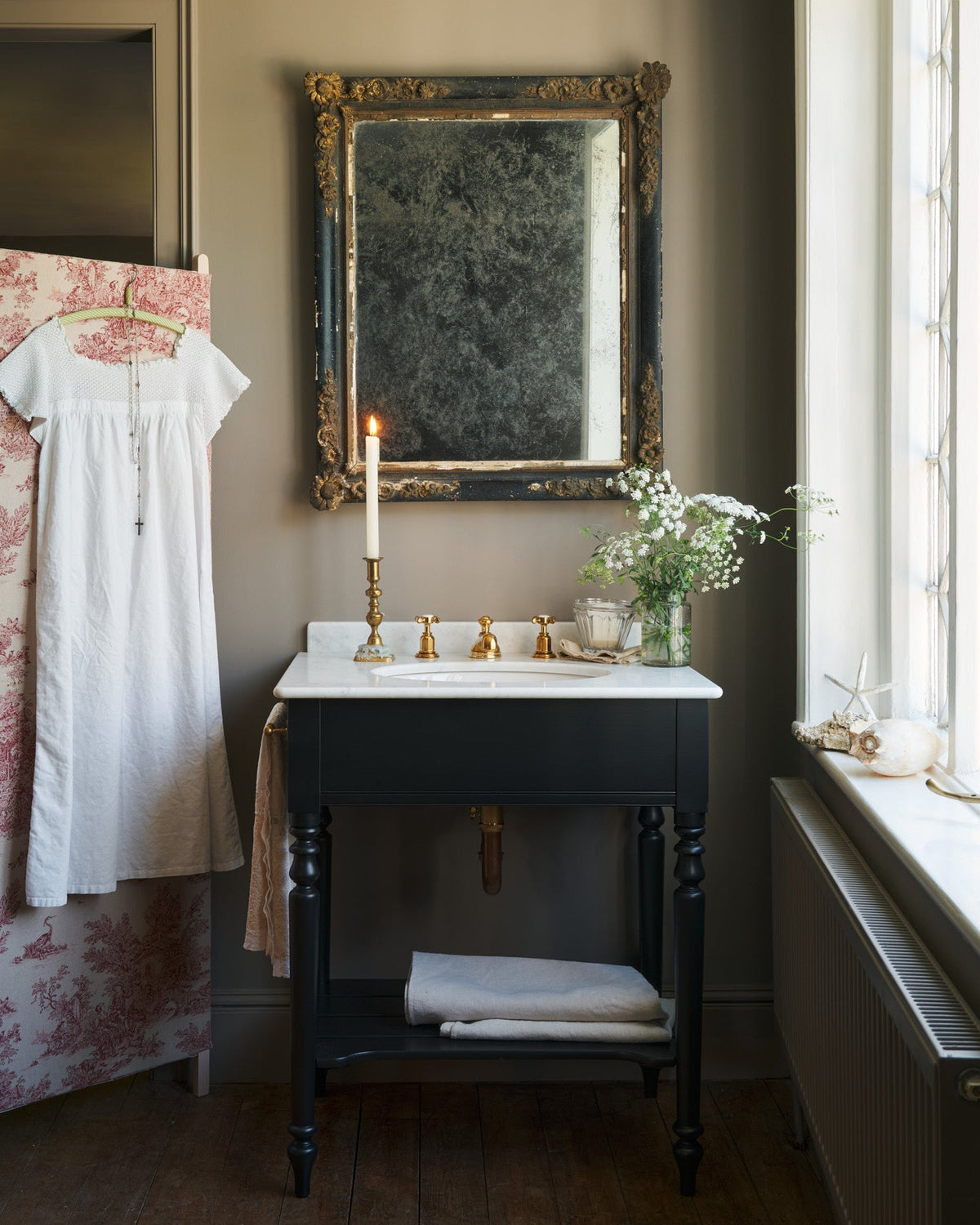 A straight-on view of a black Victorian-inspired washstand with a brass mixer tap in a greyish-brown painted room. A mottled antique mirror sits above the sink, an antique cotton dress hangs from a decorated partition and light streams through a nearby leaded window.