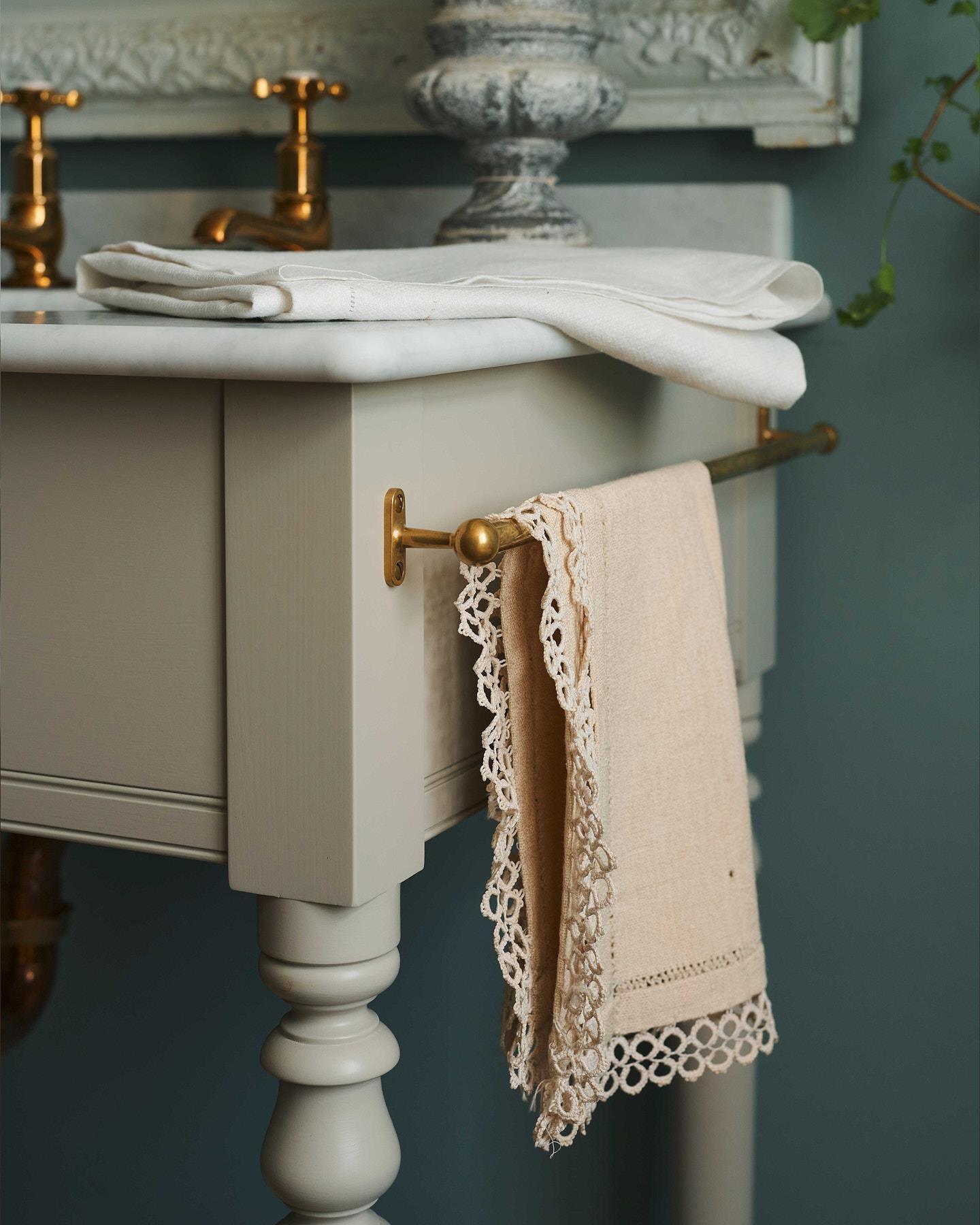 A detailed view of an aged brass towel rail attached to the right side of a Victorian-inspired washstand, a vintage lace hand towel hangs from the rail.