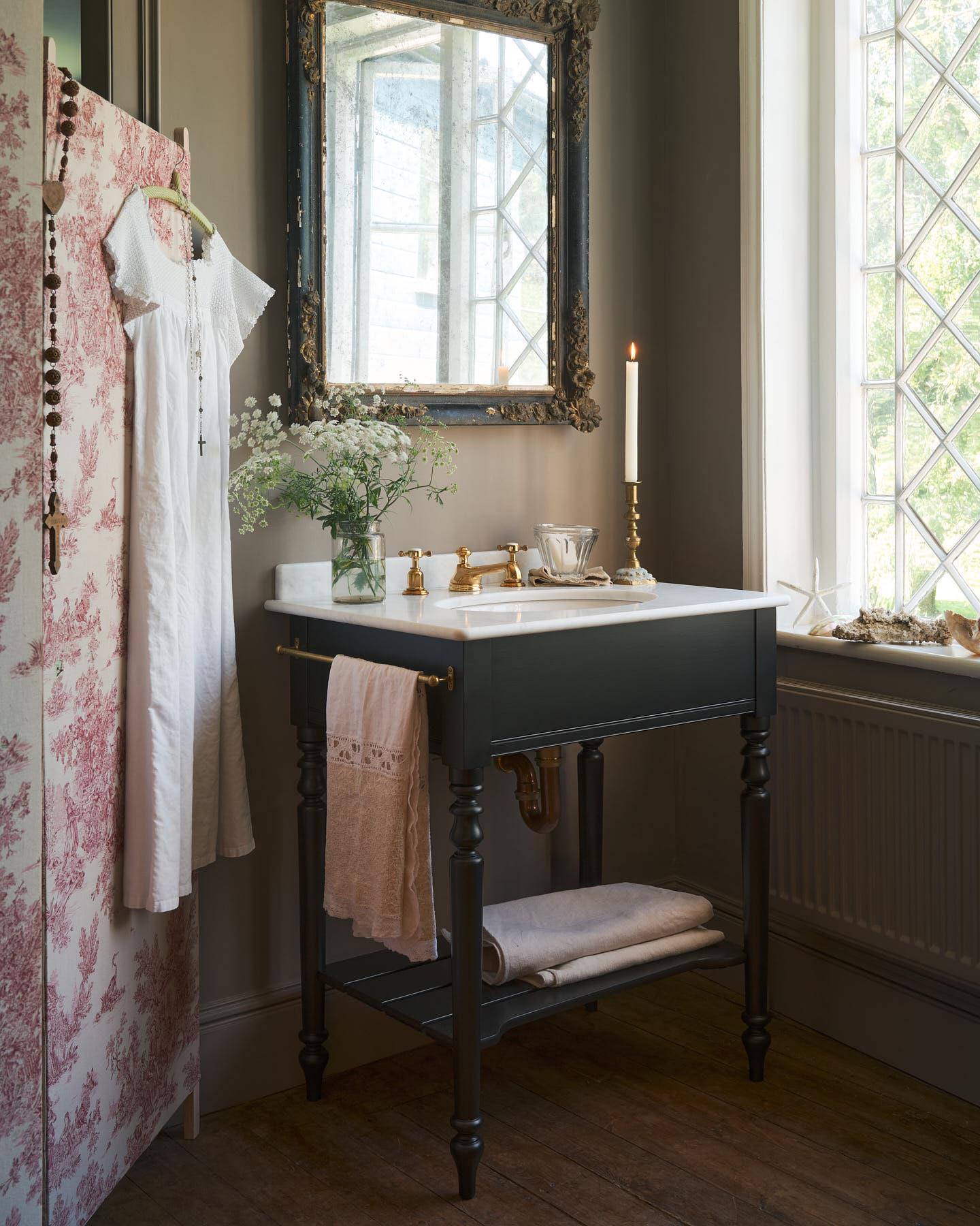 An oblique view of a black Victorian-inspired washstand with a brass mixer tap in a greyish-brown painted room. A mottled antique mirror sits above the sink, an antique cotton dress hangs from a decorated partition and light streams through a nearby leaded window.