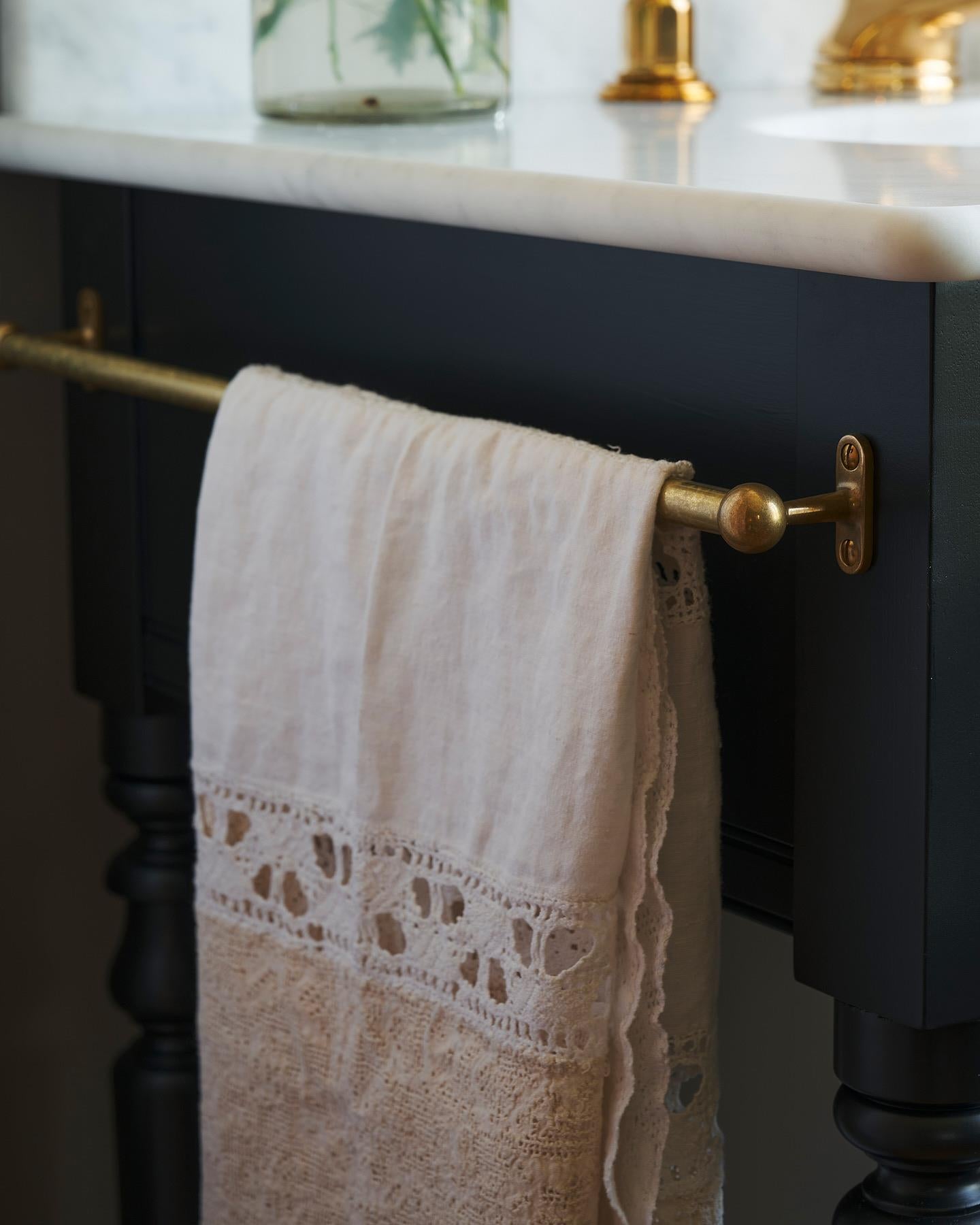 A detailed view of an aged brass towel rail attached to the left side of a Victorian-inspired washstand, a vintage lace hand towel hangs from the rail.