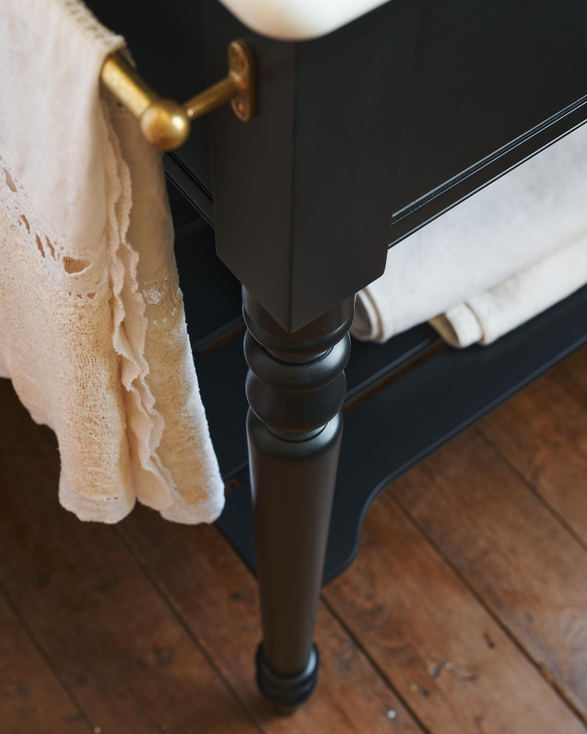A detailed view of an aged brass towel rail attached to the left side of a Victorian-inspired washstand, a vintage lace hand towel hangs from the rail. TEST