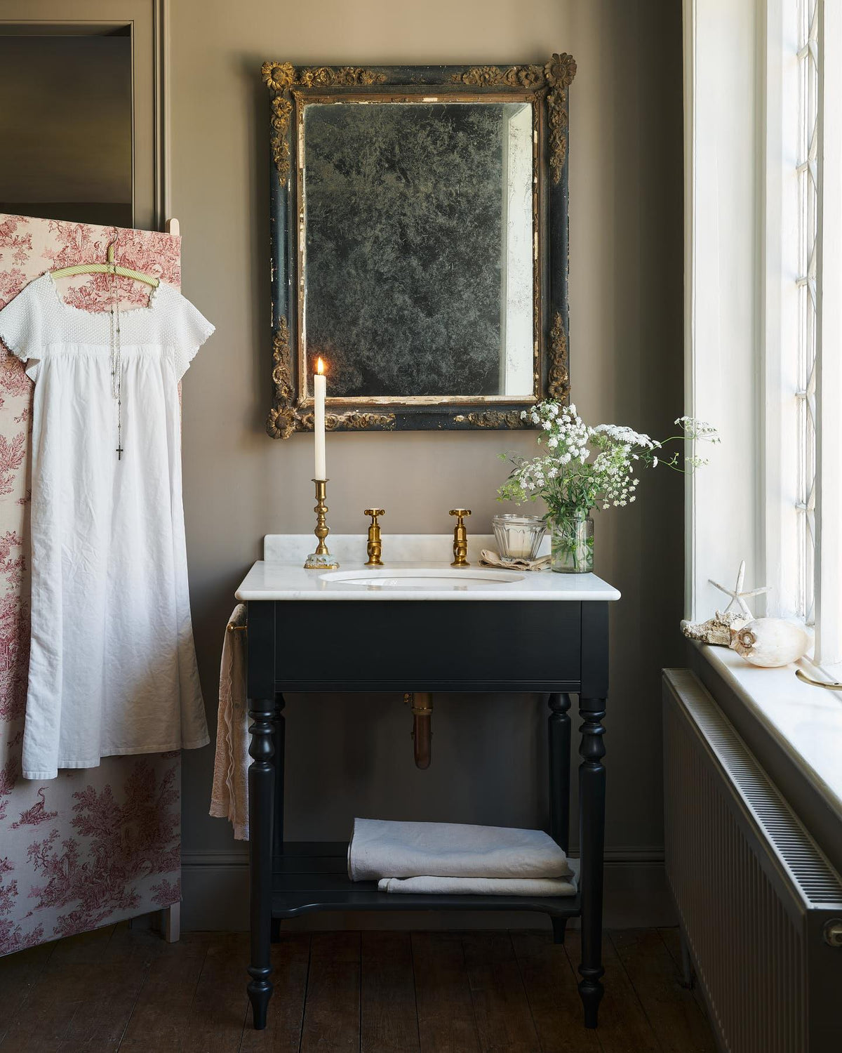 A straight-on view of a black Victorian-inspired washstand with a brass pillar taps in a greyish-brown painted room. A mottled antique mirror sits above the sink, an antique cotton dress hangs from a decorated partition and light streams through a nearby leaded window. | Medium Printer's Black shown TEST