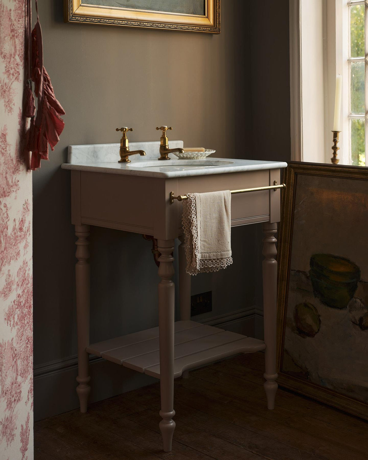 An oblique view of a dusty-pink Victorian-inspired washstand with brass pillar taps in a greyish-brown painted room. The shot is atmospheric and moody, a vintage lace towel hangs from a brass rail attached to the front of the washstand. TEST