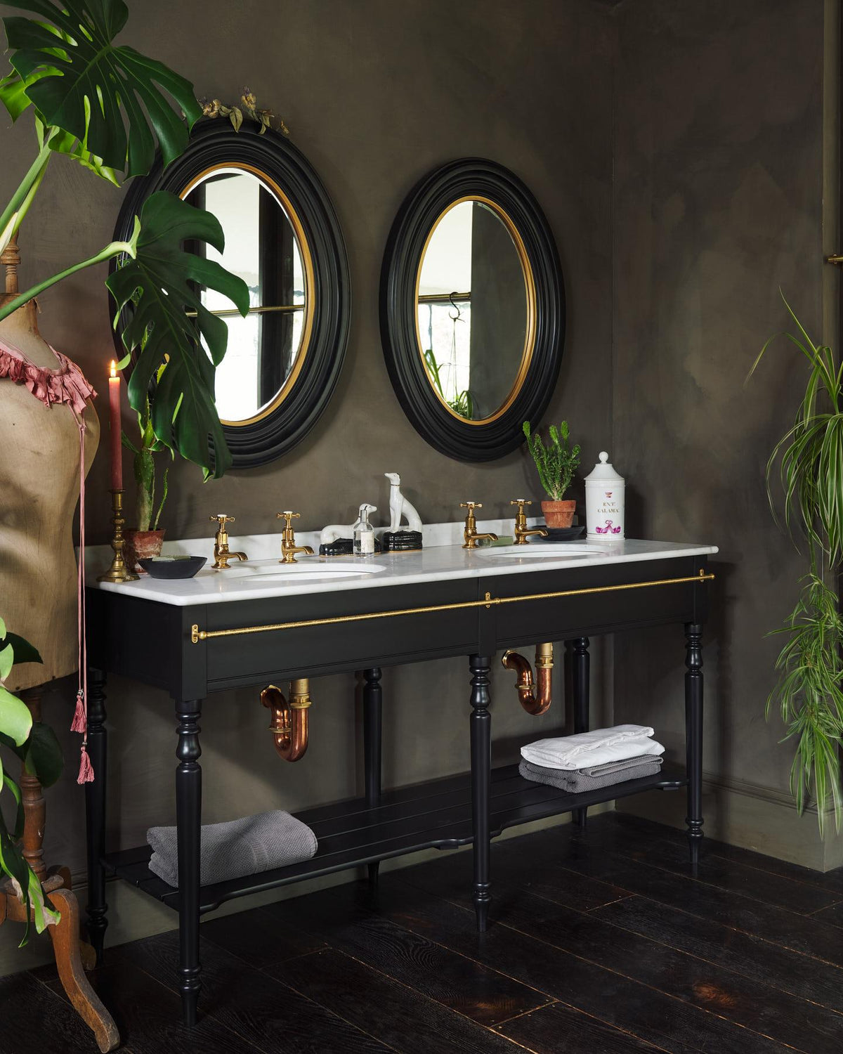 An oblique view of a grand black Victorian-inspired washstand with two basins and two sets of brass pillar taps. Two black-framed oval mirrors sit above the basins, beside a large monstera plant and eclectic styling with vintage bric-a-brac. | Printer's Black shown TEST