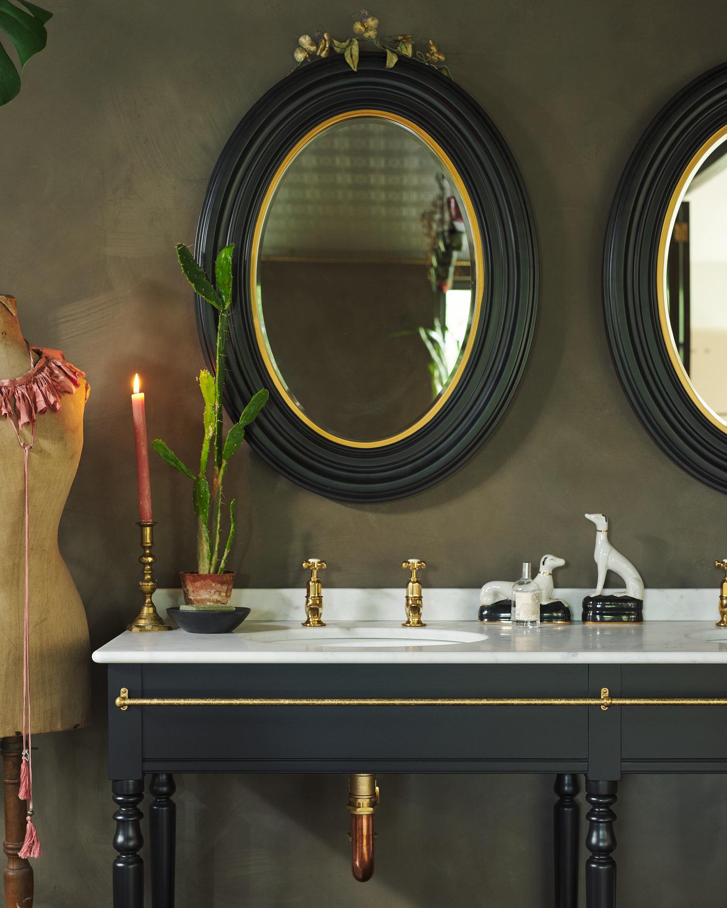 A close-up, straight-on view of a grand black Victorian-inspired washstand with two basins and two sets of brass pillar taps. Two black-framed oval mirrors sit above the basins, beside a large monstera plant and eclectic styling with vintage bric-a-brac.