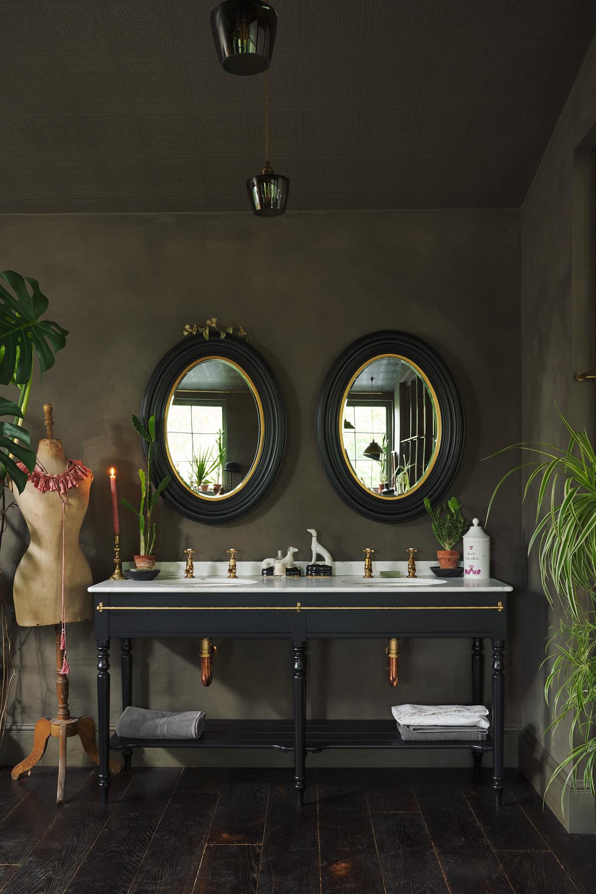 A straight-on view of a grand black Victorian-inspired washstand with two basins and two sets of brass pillar taps. Two black-framed oval mirrors sit above the basins, beside a large monstera plant and eclectic styling with vintage bric-a-brac. | Pillar Taps shown TEST