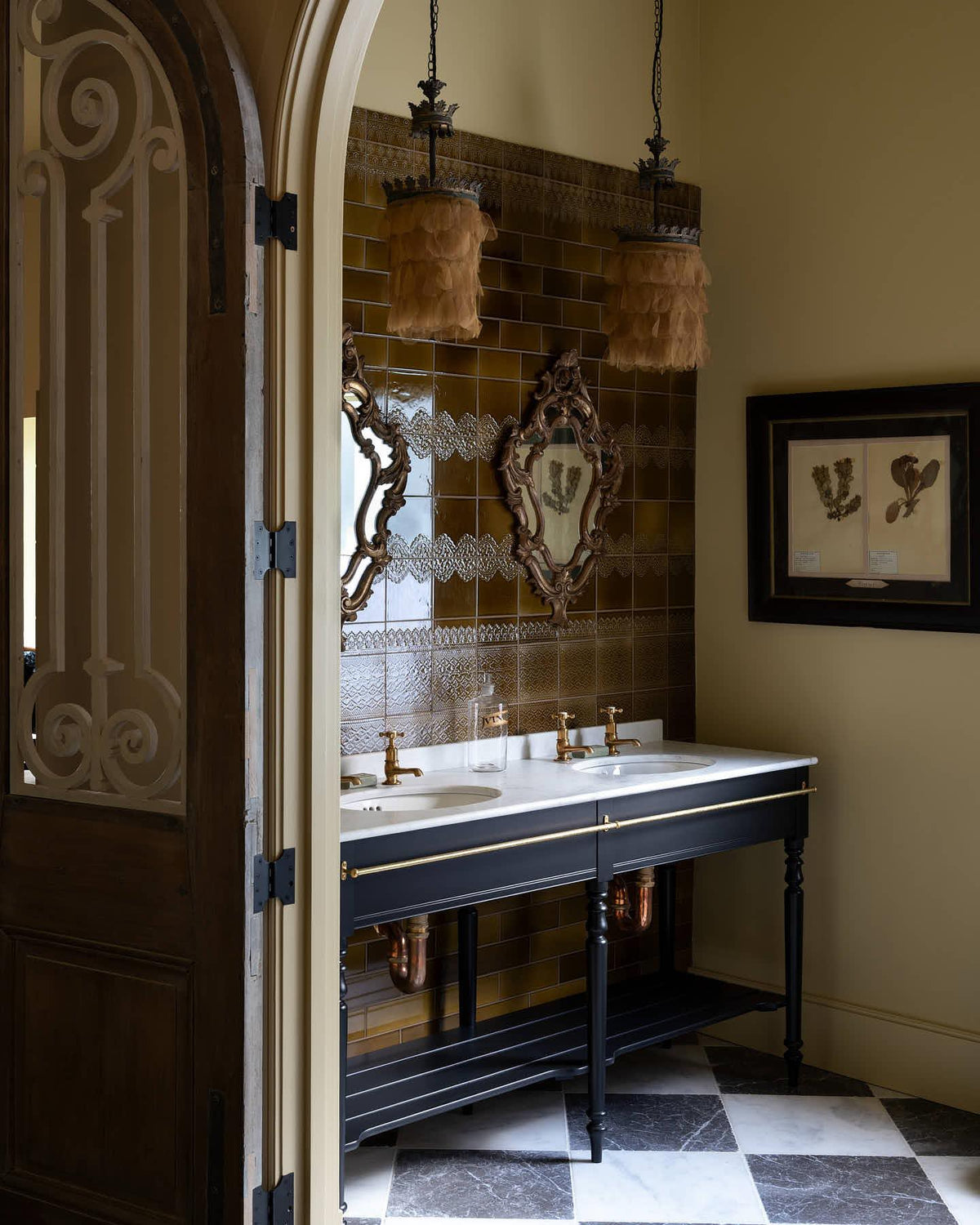 An oblique view of a grand black Victorian-inspired washstand with two basins and two sets of brass pillar taps. Two antique mirrors and pendant lights sits above the basins. All set against a tall wall of sepia lace market tiles. | Printer's Black shown TEST